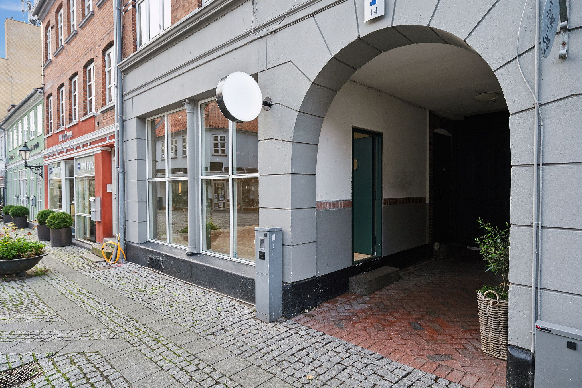 Bygning med en stor buegang og grøn dør, der fører ind til en parkeringsplads. Bag bygningen er der en butik med røde vægge