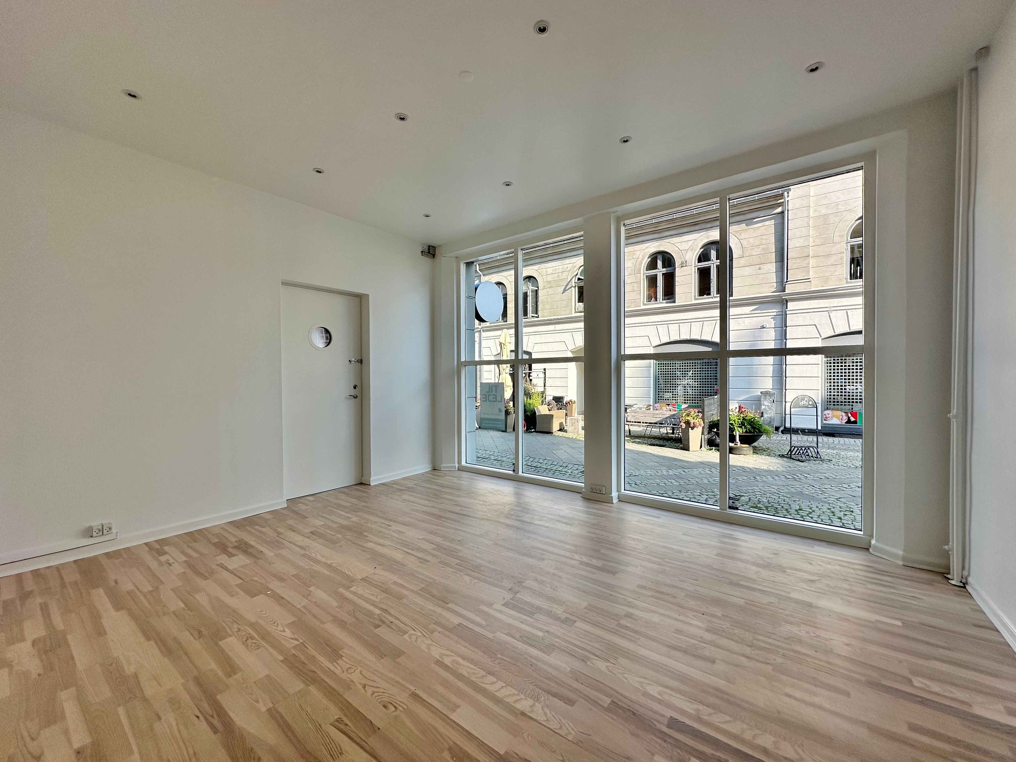 Store glasruder med udsigt til gågaden, lyse lofter, hvide vægge og trægulv. I udsigten er der en stor bygning med flere