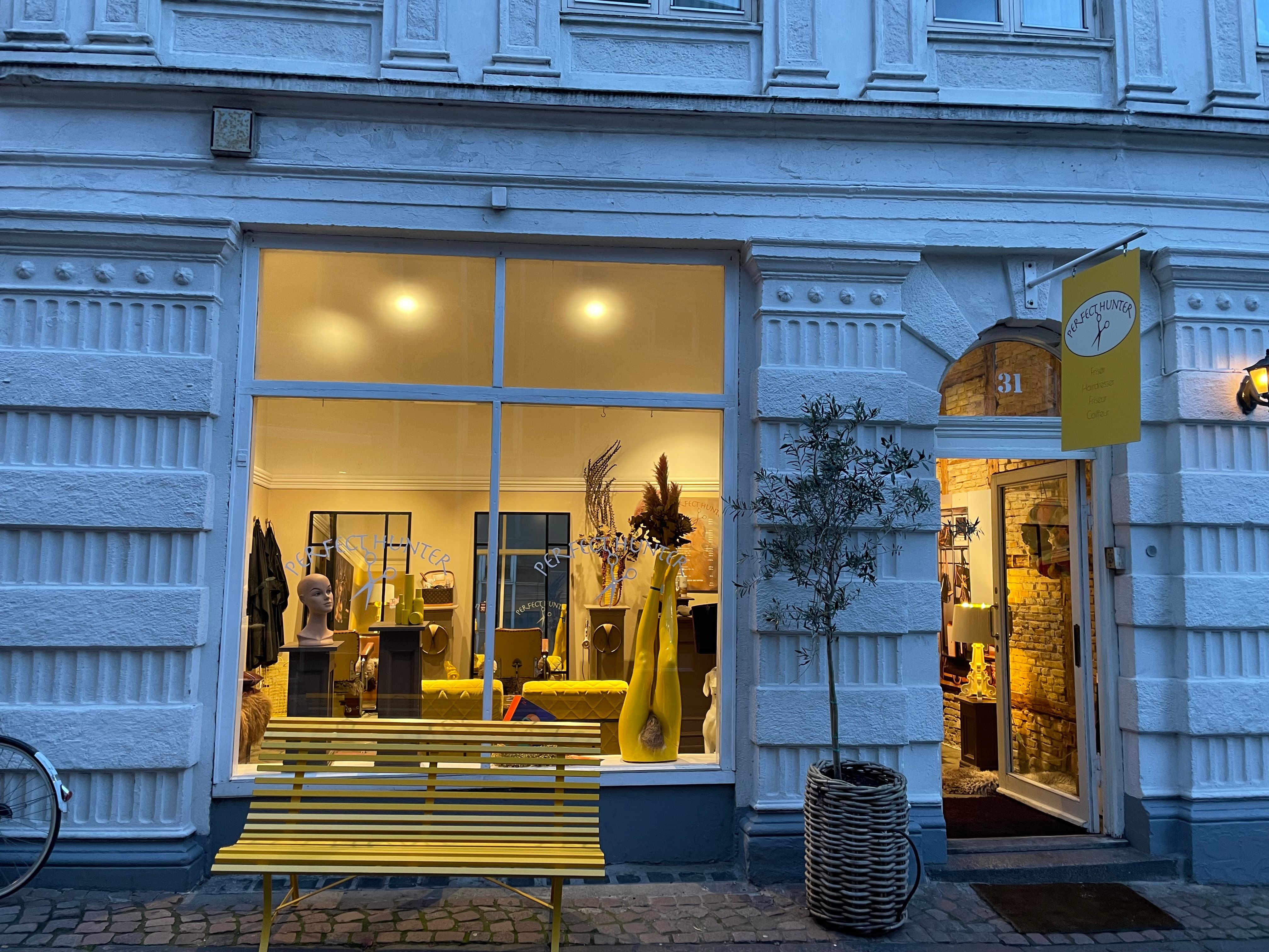 Lys og smukke butiksvinduer med lysegul træbænk og oliventræ. Lyserød banner med teksten "Perfect Hunter" og nummer 31.