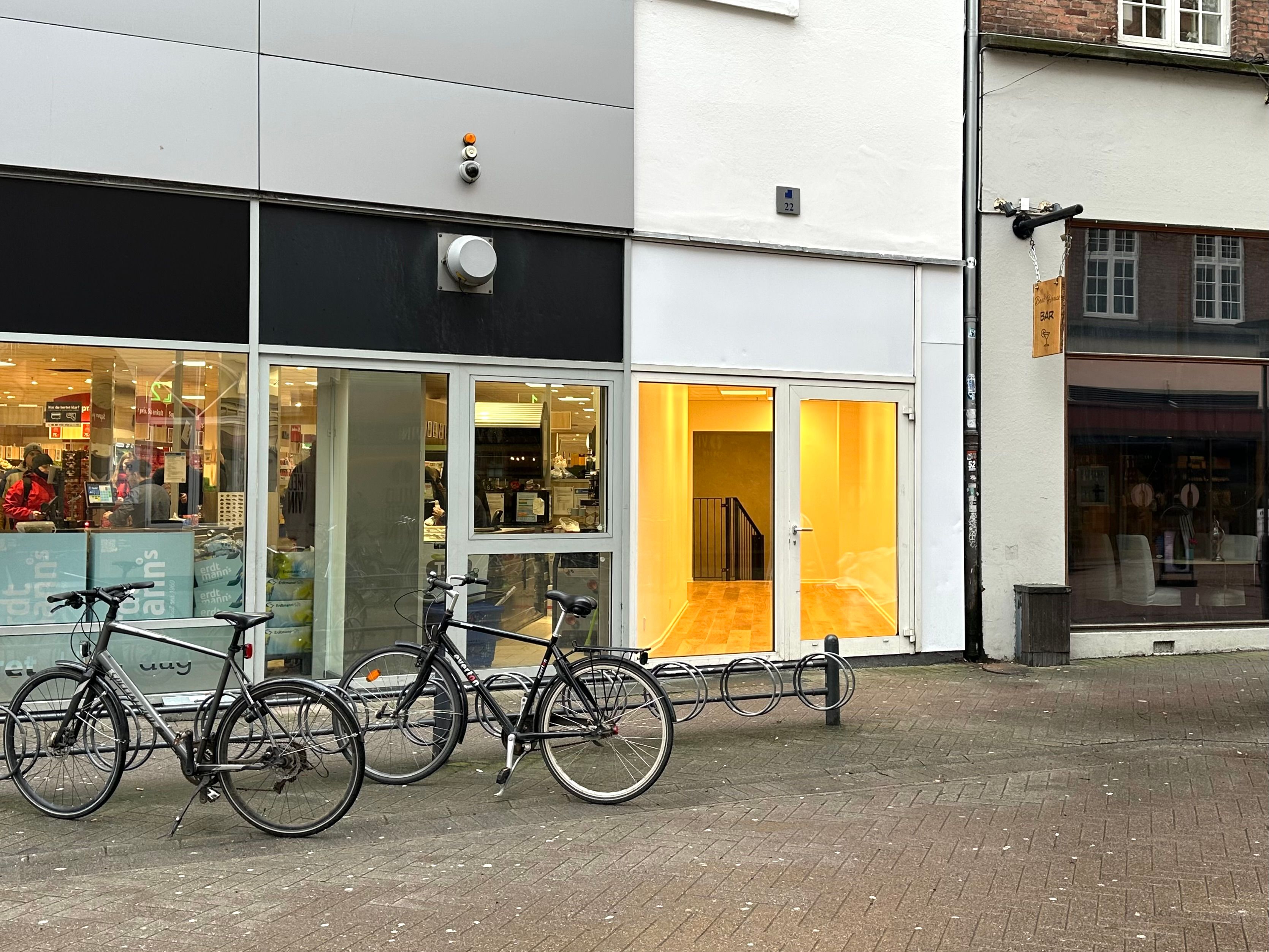 Butikker med glasruder og åbne døre på Slotsgade. Tre cykler står parkeret udenfor og kameraer er monteret på bygningerne. Et schild med tekst hænger udenfor den højre bygning.