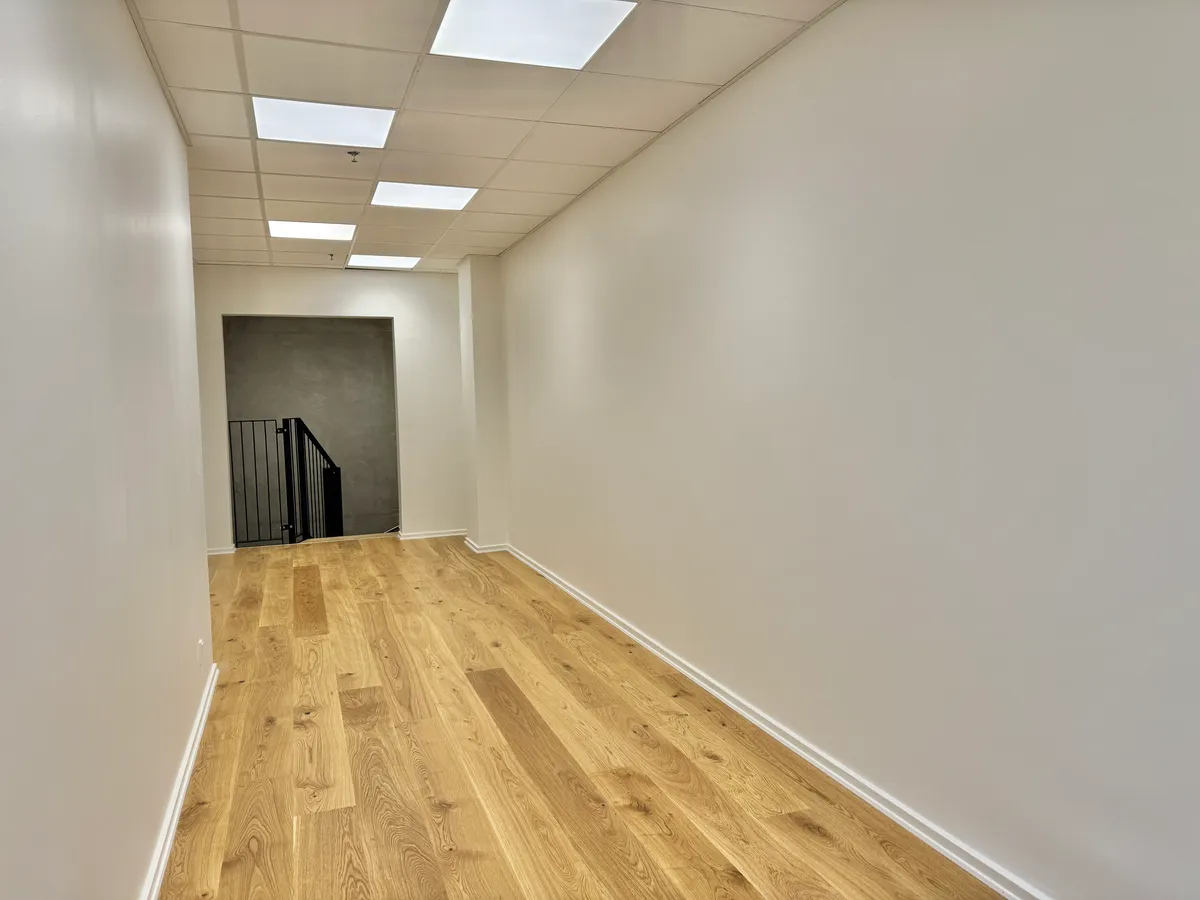 Indersiden af lejlighed med lyse loftslamper, hvidt malet væg og trægulv og åbning til trappeopgang med metalgelænder