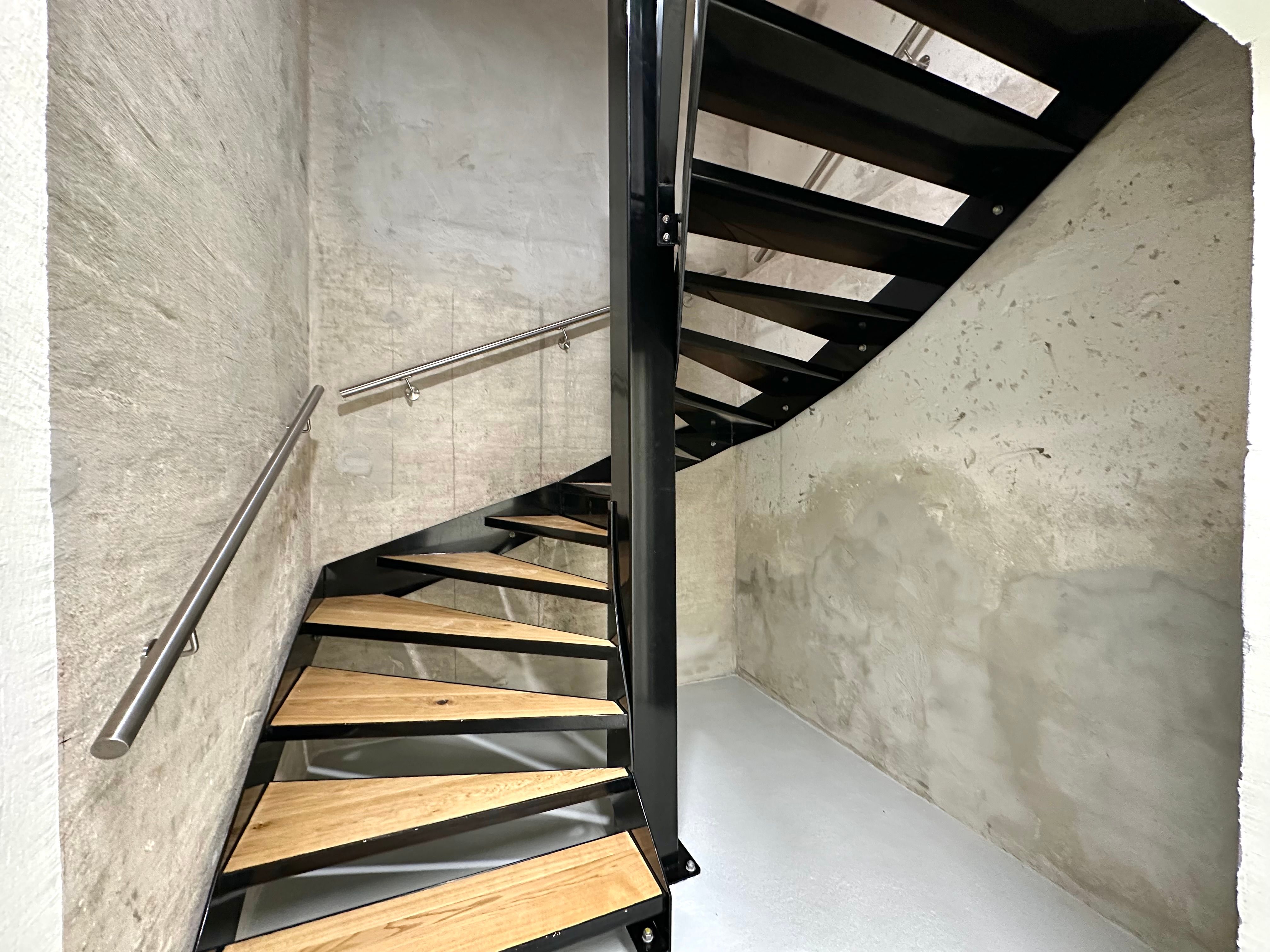 Elegant skrå trætrappe med sort metalgehus og skinnende stålhåndtag, som fører op til første sal i en ombygget bygning med hvid og grå vægbeklædning