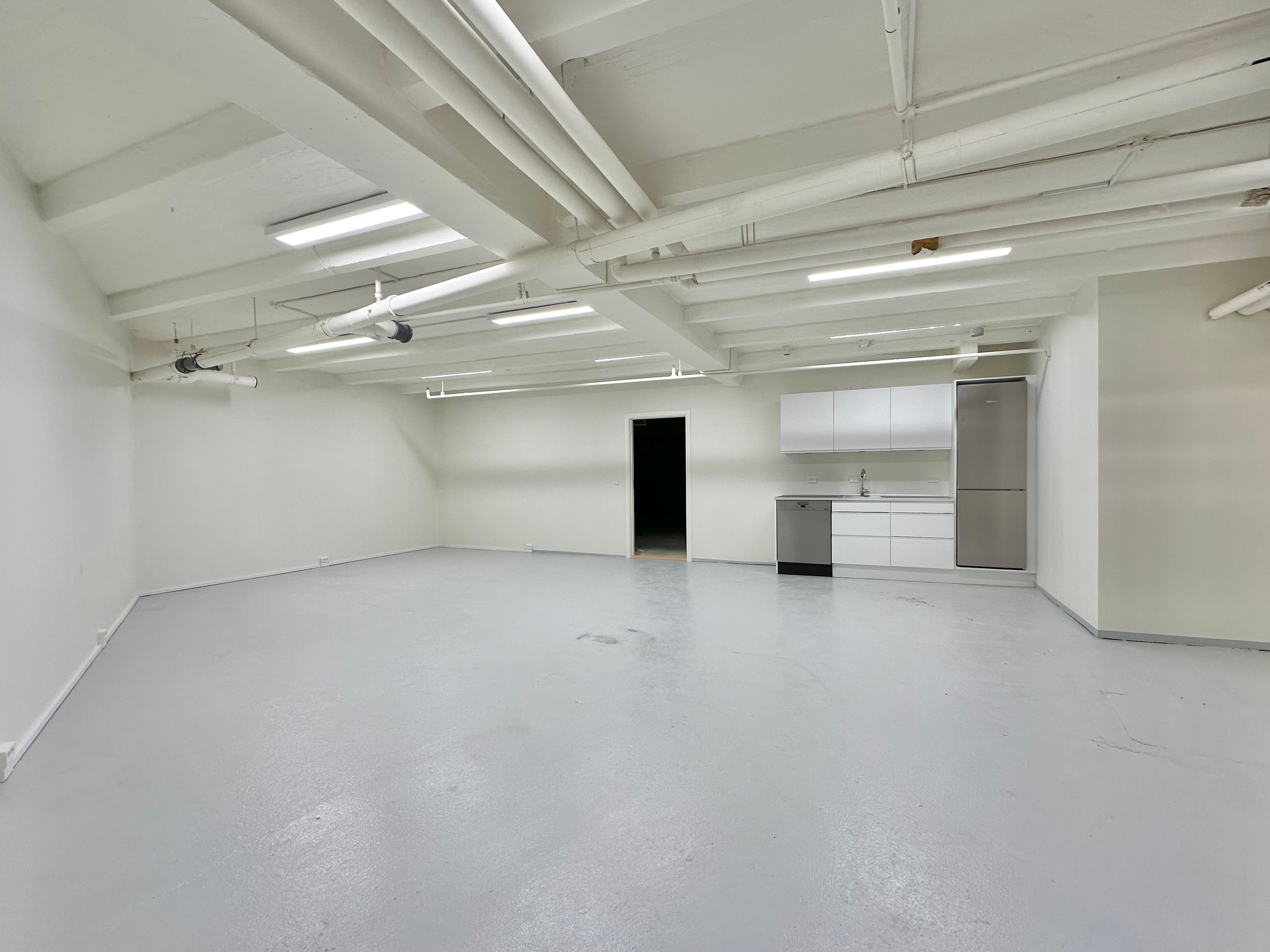 Et stort hvidt lokale med en køkkendel, hvor der er en vaskemaskine og køleskab, høje lofter og lyse armaturer