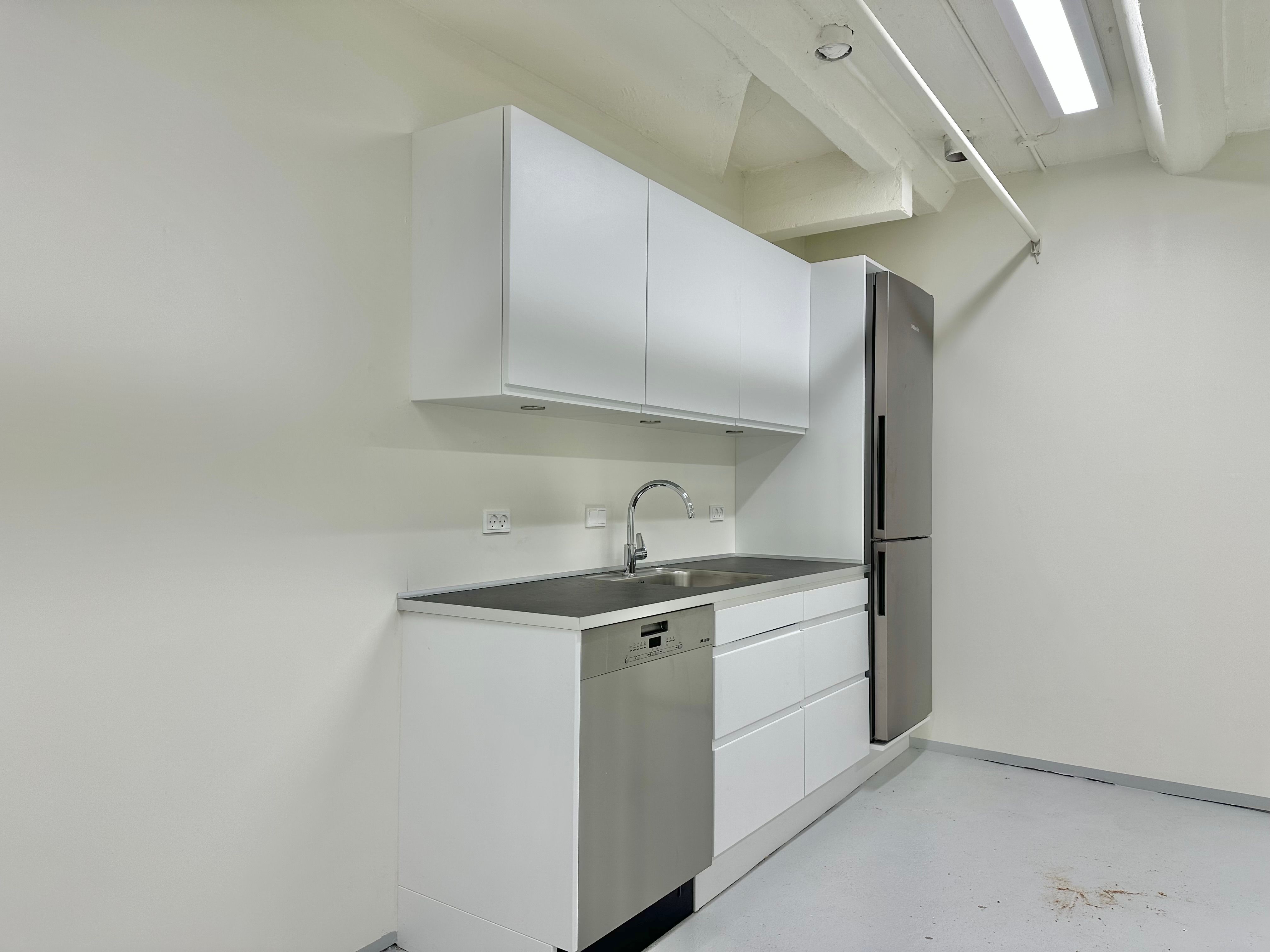 Rummelig kælder med moderne køkken. Hvidt køkken med højloft, rustfrit stålkøleskab og indbygget vaskemaskine. To hvide skabe med to hængehylder. Køkken med en vandhane og trægulv