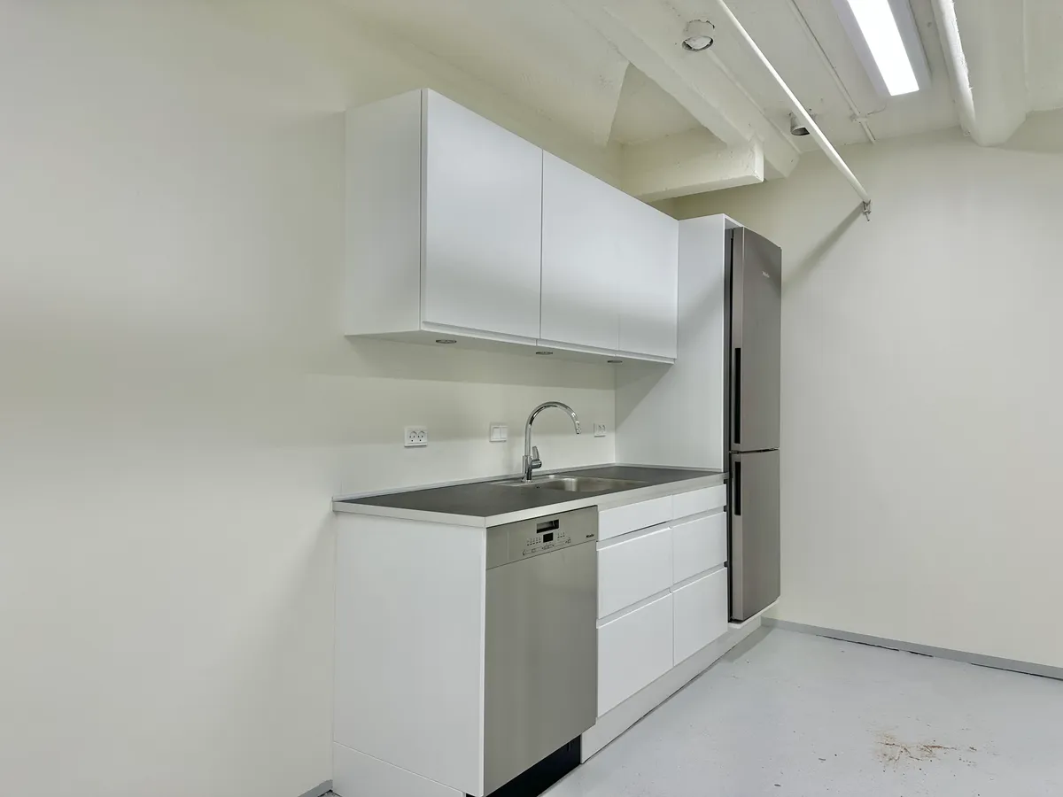 Rummelig kælder med moderne køkken. Hvidt køkken med højloft, rustfrit stålkøleskab og indbygget vaskemaskine. To hvide skabe med to hængehylder. Køkken med en vandhane og trægulv