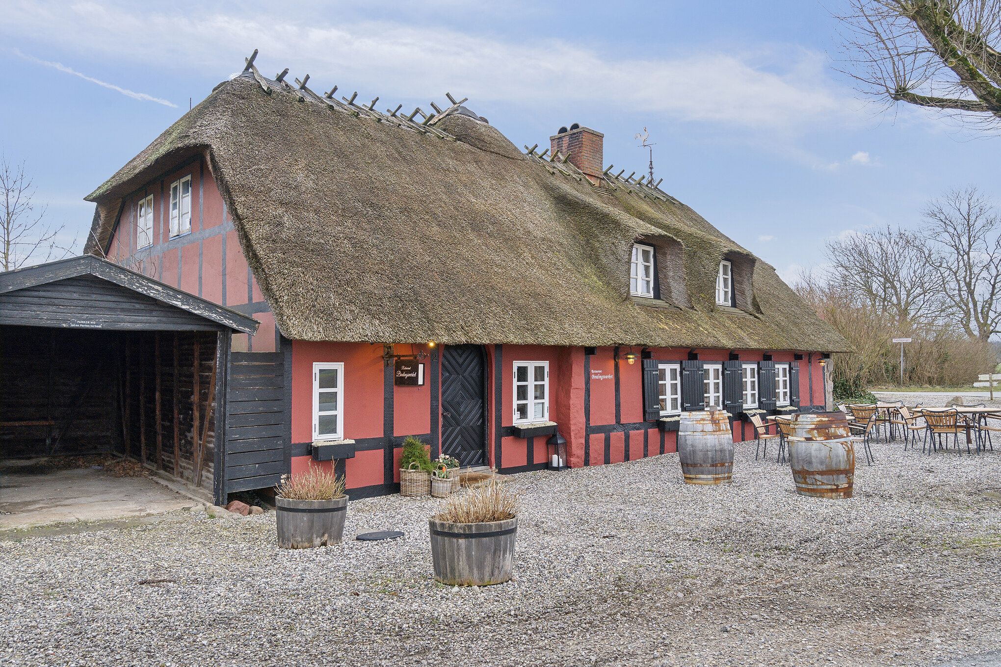 Rødt bindingsværkshus med røgkammer, halmtag og små vinduer. Udsmykket med lyserøde potter og træfæsthed.
