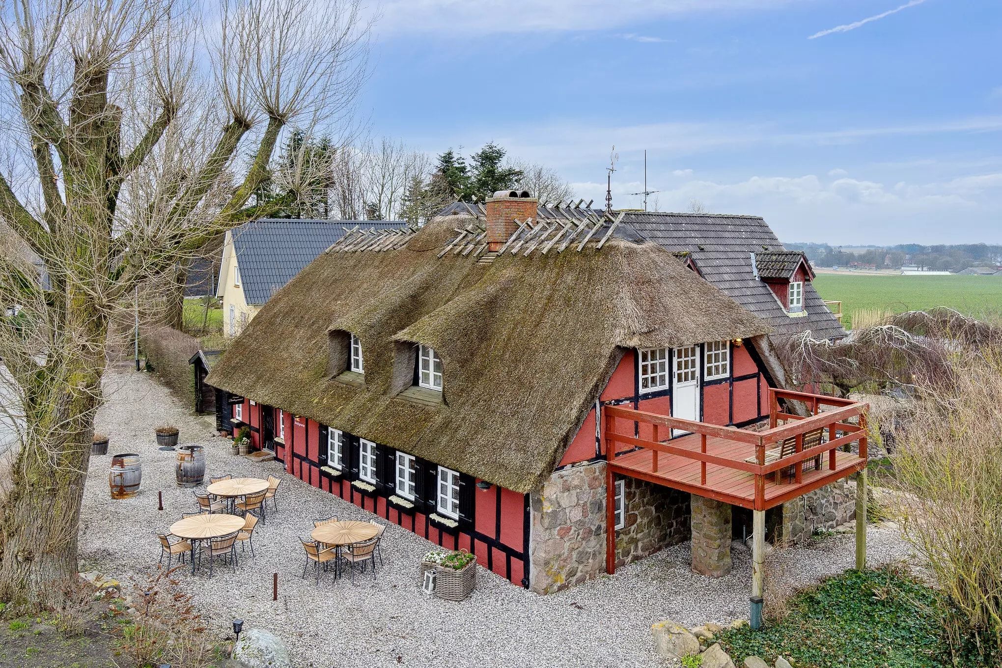 En gammel dansk gård med et rødt tækt tag og træværanda samt et række af træstole og borde