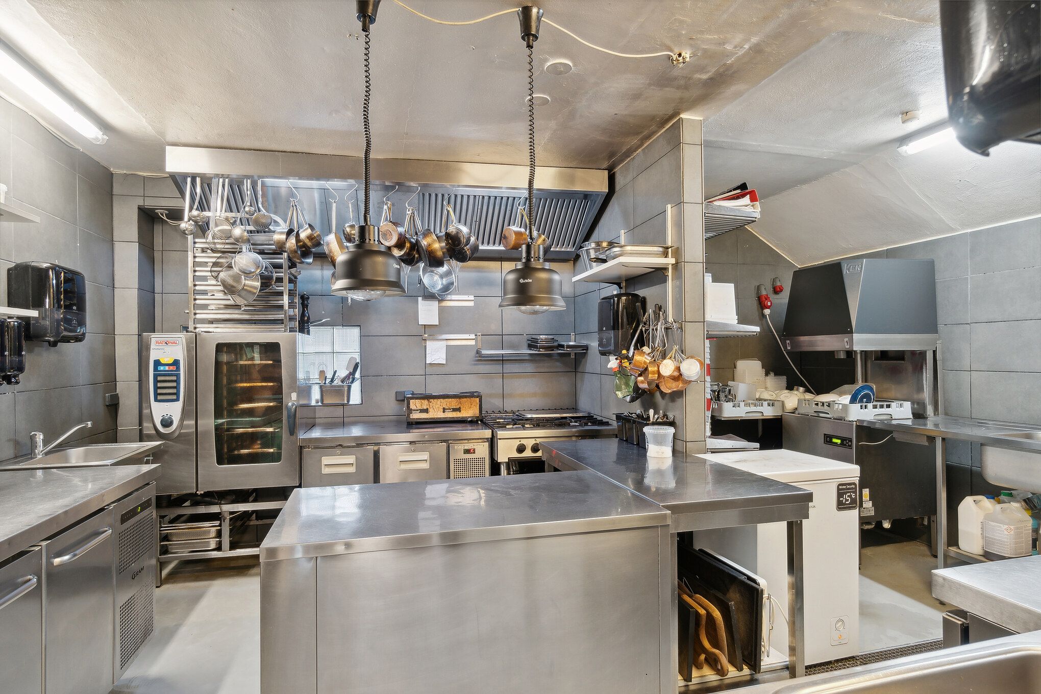En professionel køkken med to store kokepladser, ovne, fryseskabe, kogegrej og flere apparater og møbler.