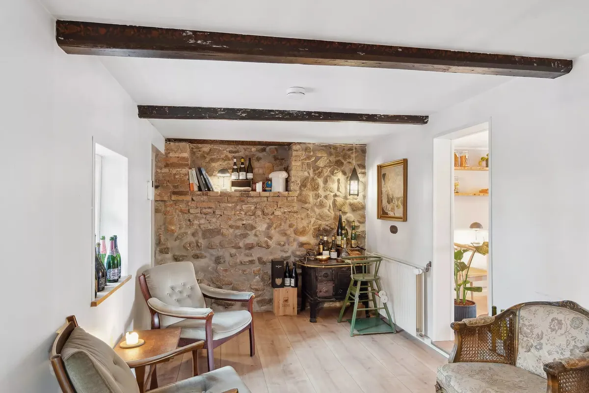 Indvendigt i et hus med hvidt vægmaleri og trægulv er der et stolsæt, et bord og en ølkøler med flasker