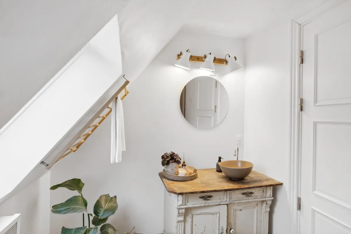 Rundt spejl i hvidt badeværelse med lysekroner, hængende håndklæde, plante, træskab og vandhane med rustfrit stål