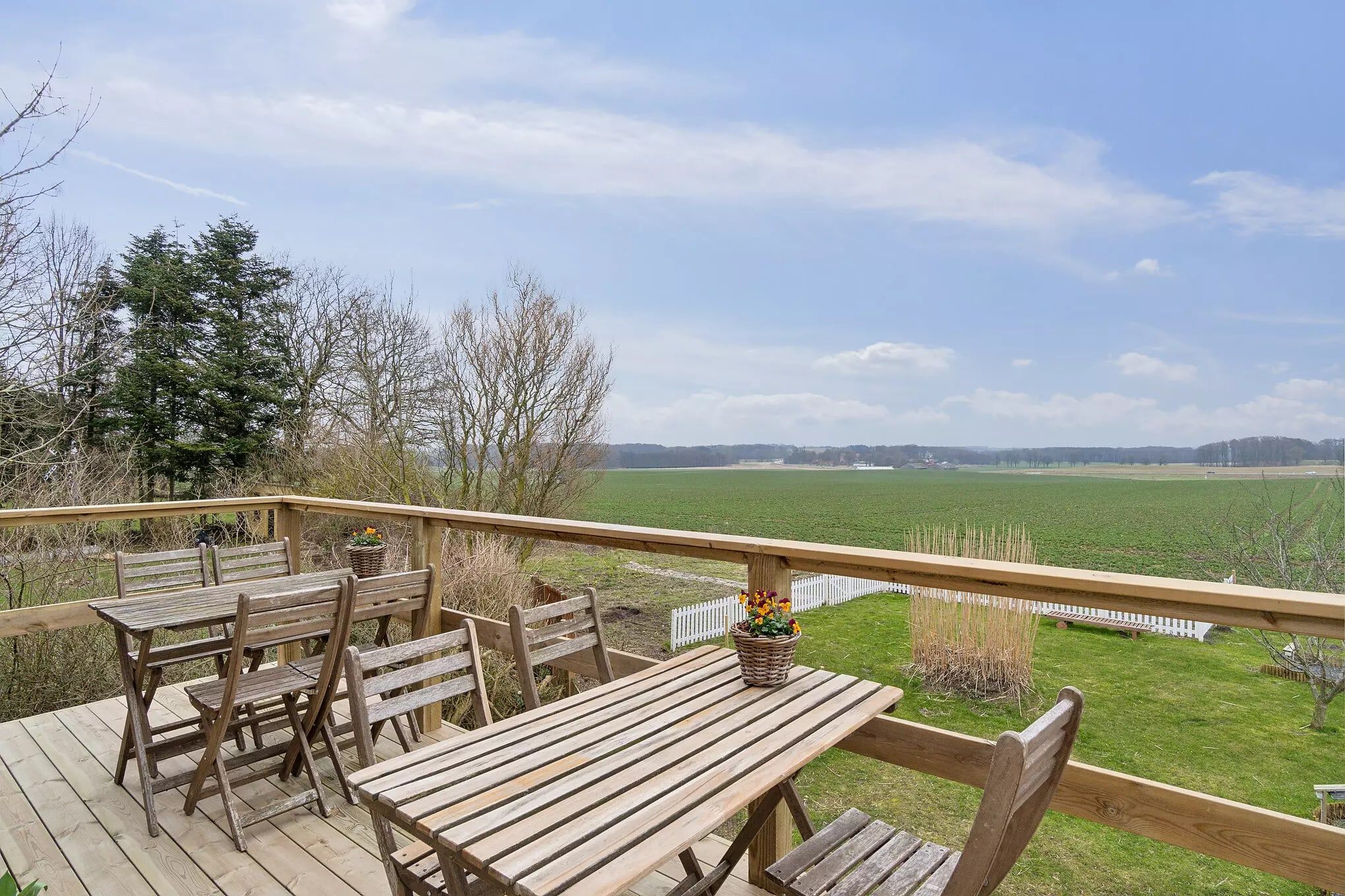 Træterrasse med udsyn til grønne marker og en hvid lav hegn, med to træborde og stole samt blomster i kurve