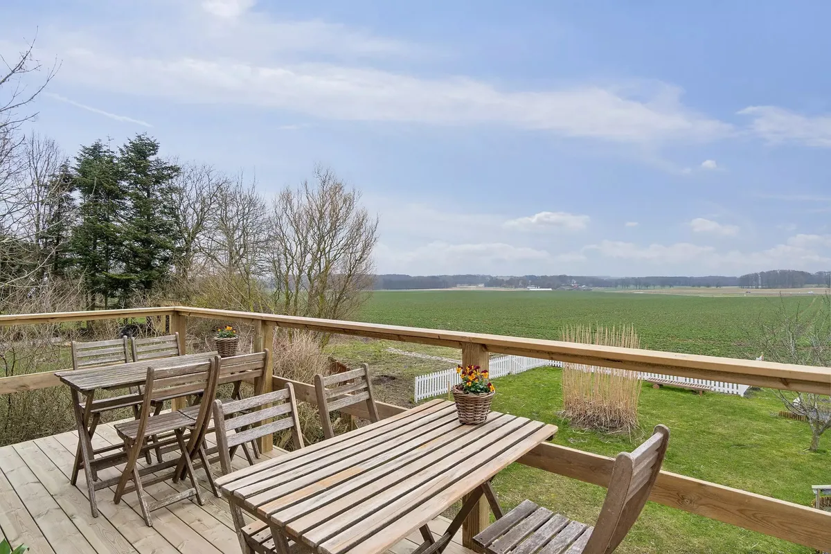 Træterrasse med udsyn til grønne marker og en hvid lav hegn, med to træborde og stole samt blomster i kurve