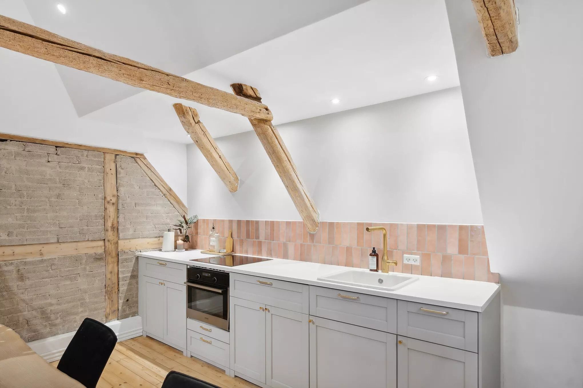 Moderne køkken med hvide skabe og loftsbjælker, hvor der er en gaskomfur, hængende puder og en væg med bakstensmønster