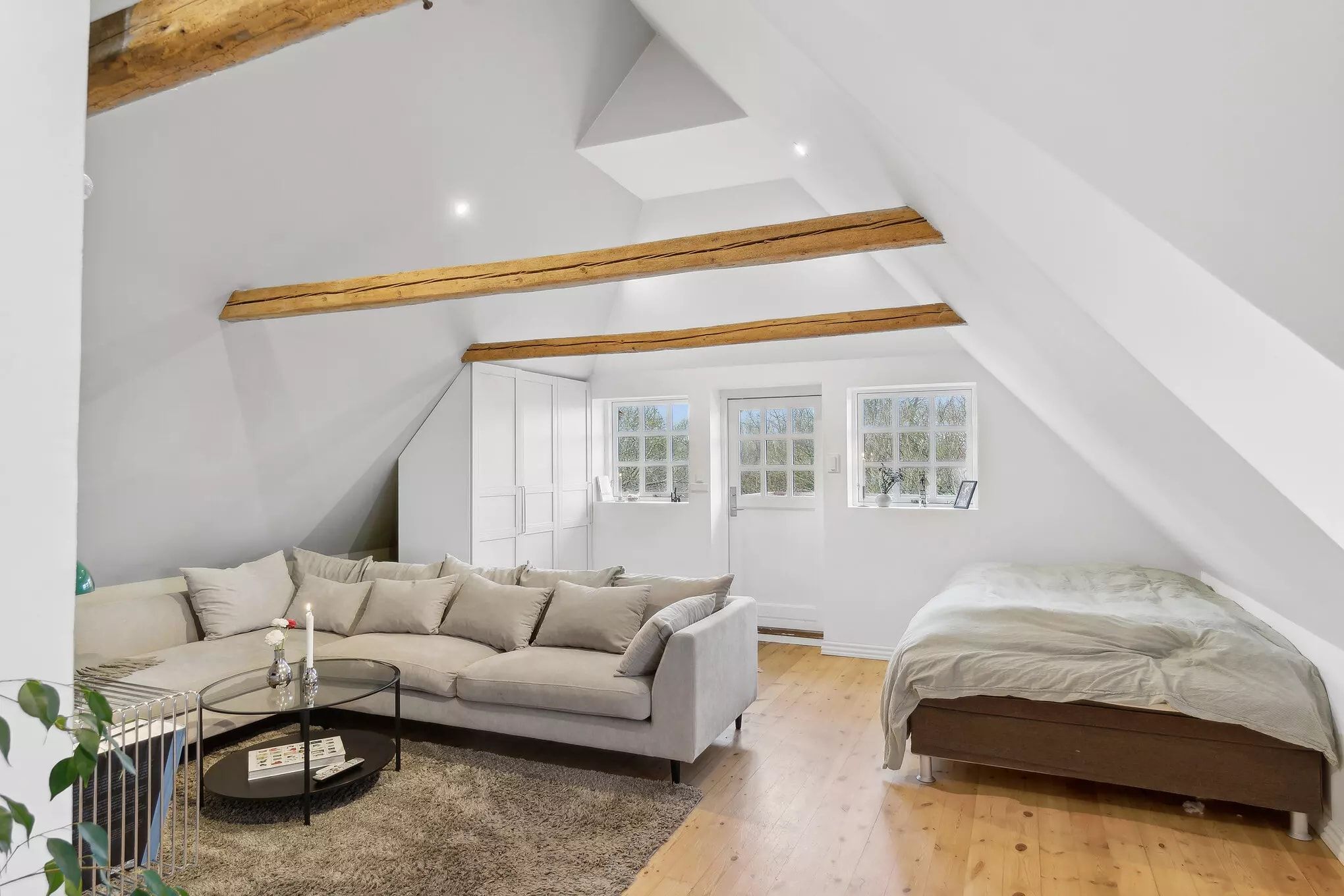 Inde i et soveværelse med hvide vægge, loftsvinduer og træbjælker samt en sengelinned og en skovl med puder