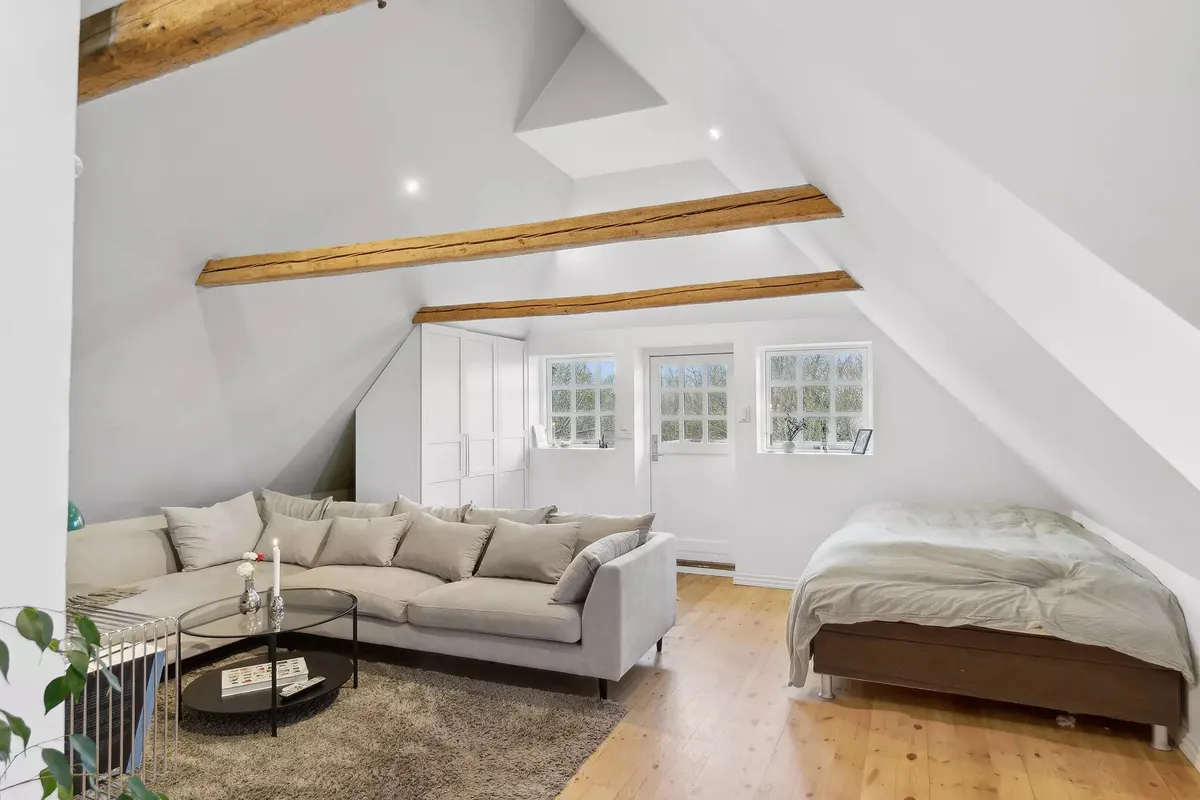 Inde i et soveværelse med hvide vægge, loftsvinduer og træbjælker samt en sengelinned og en skovl med puder