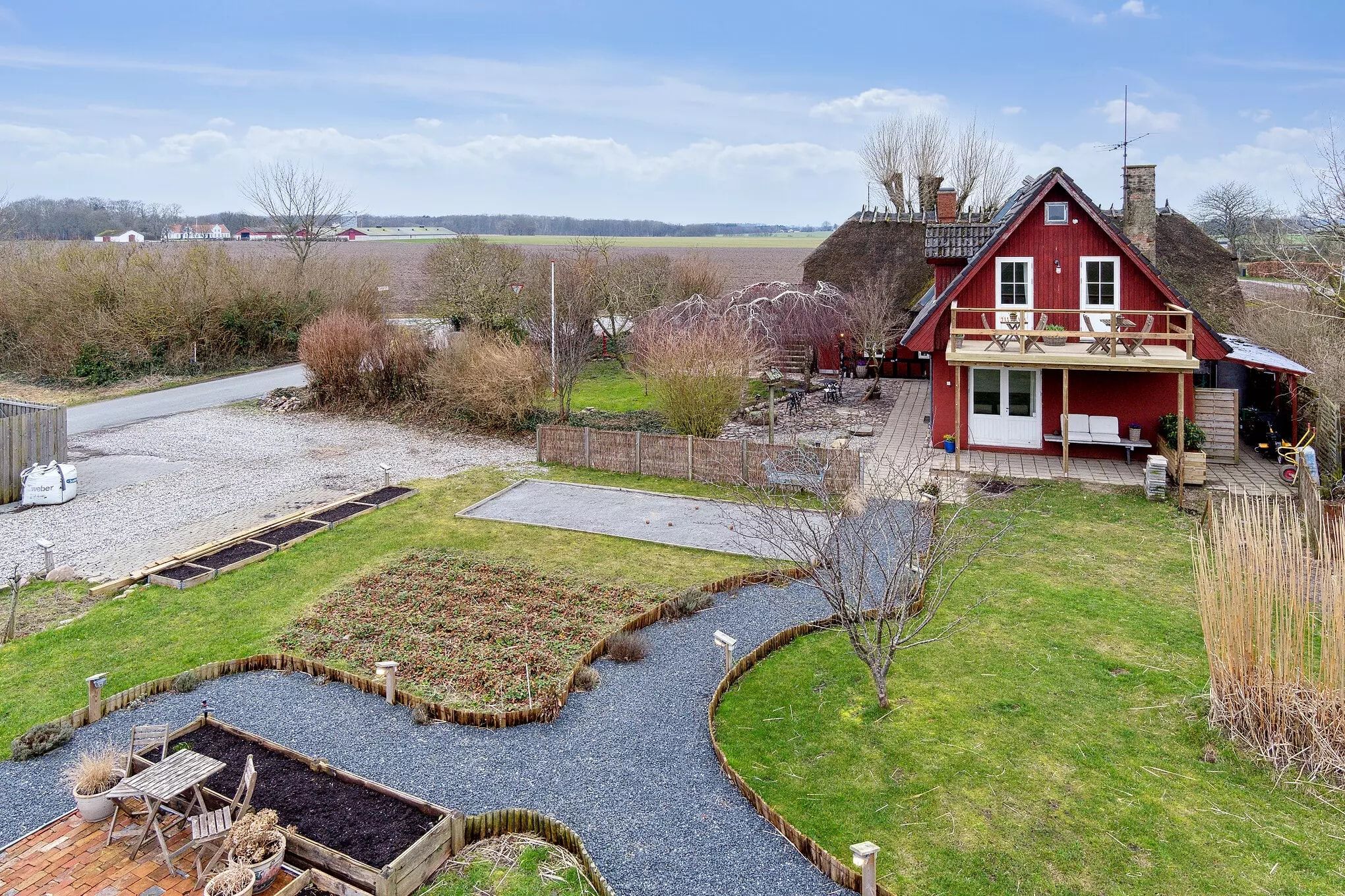 En stor rødmalet gård med en terrasse og en have med brændenælde og blomsterpotte samt en række små rækker med havebede