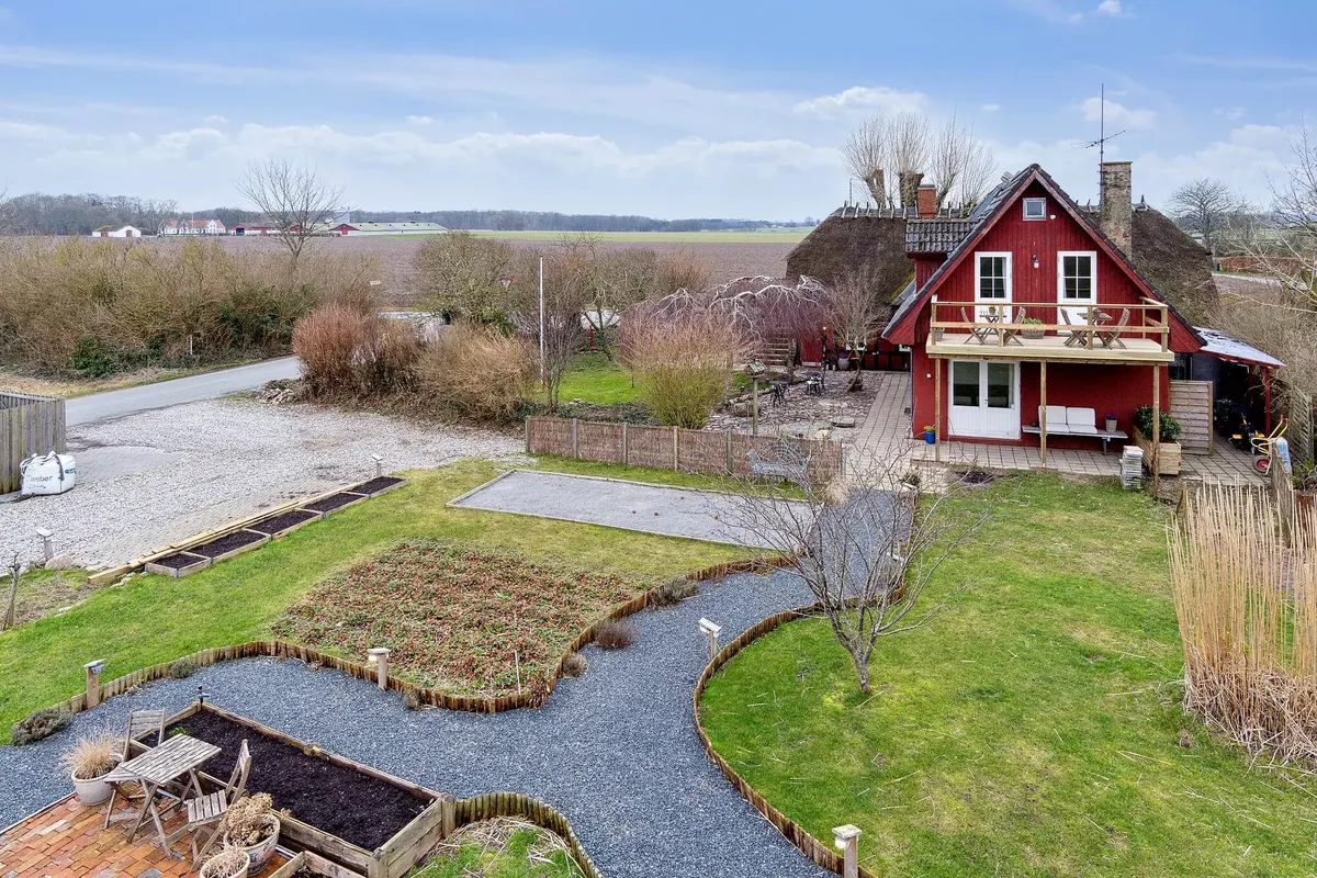 En stor rødmalet gård med en terrasse og en have med brændenælde og blomsterpotte samt en række små rækker med havebede