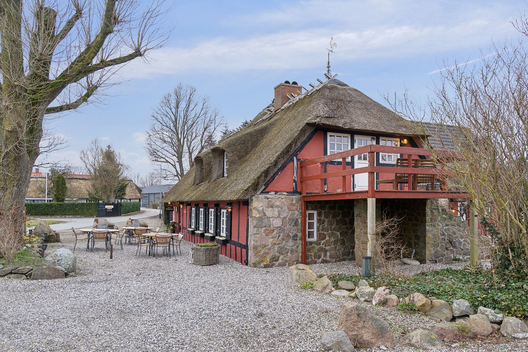Dansk landskab med rødt stenhus med stråtag og terrasse med udendørs spisning og sofaer. Bygningen har røde vægge og hvide