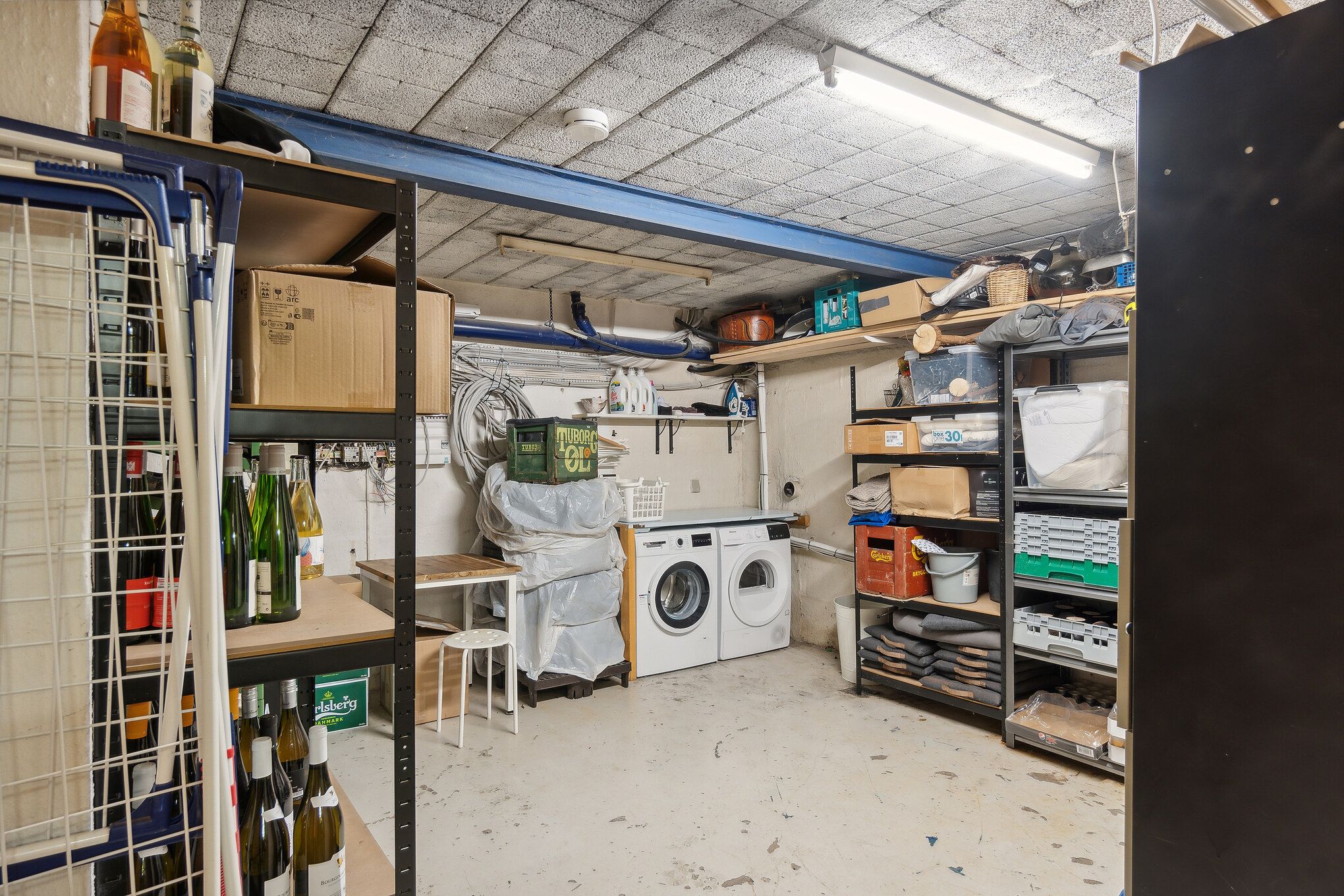 Rum med flere hylder og lagerreoler, to vaskemaskiner og en pakke tøj i vaskekurv, hvor gulvet er snavset