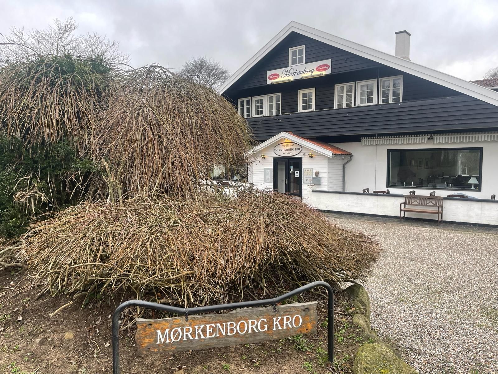 Vejledningsskilt med Mørkenborg Kro, der står i et område med tørre planter, sten og snavsede veje