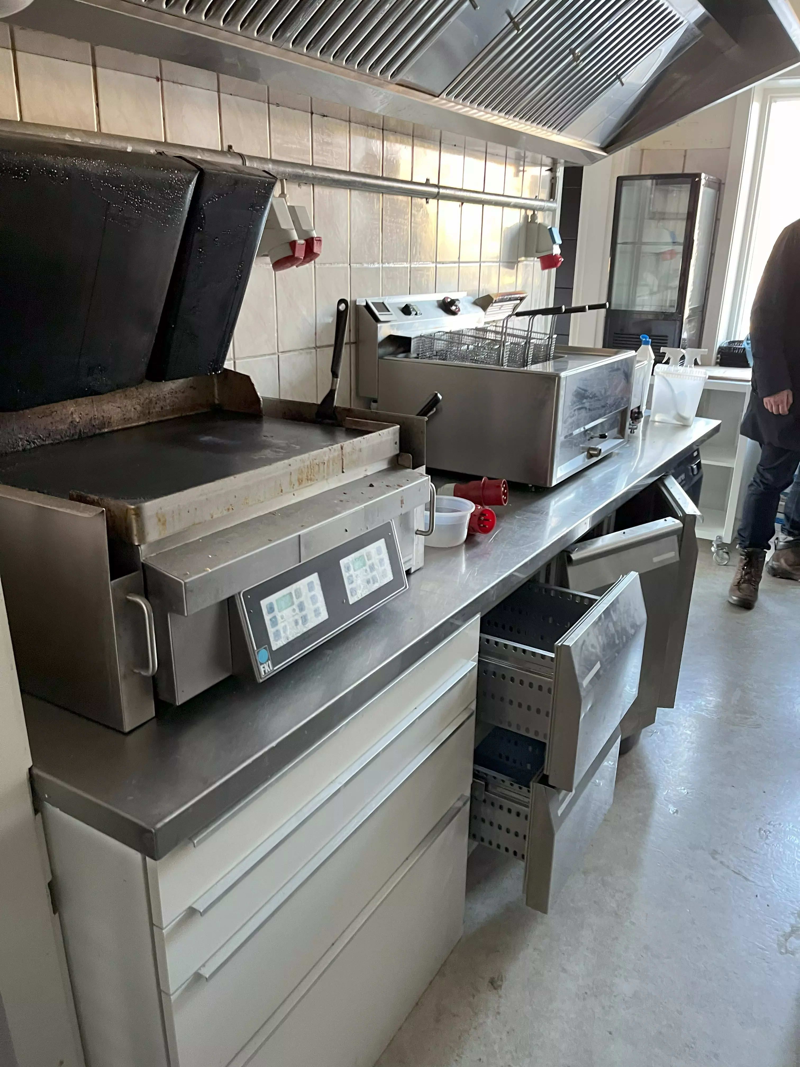 Burgerbar med moderne køkkenudstyr som grill og fritureske. Væggene er udstyret med ventiler og en stor vinduesglas. Gulvet