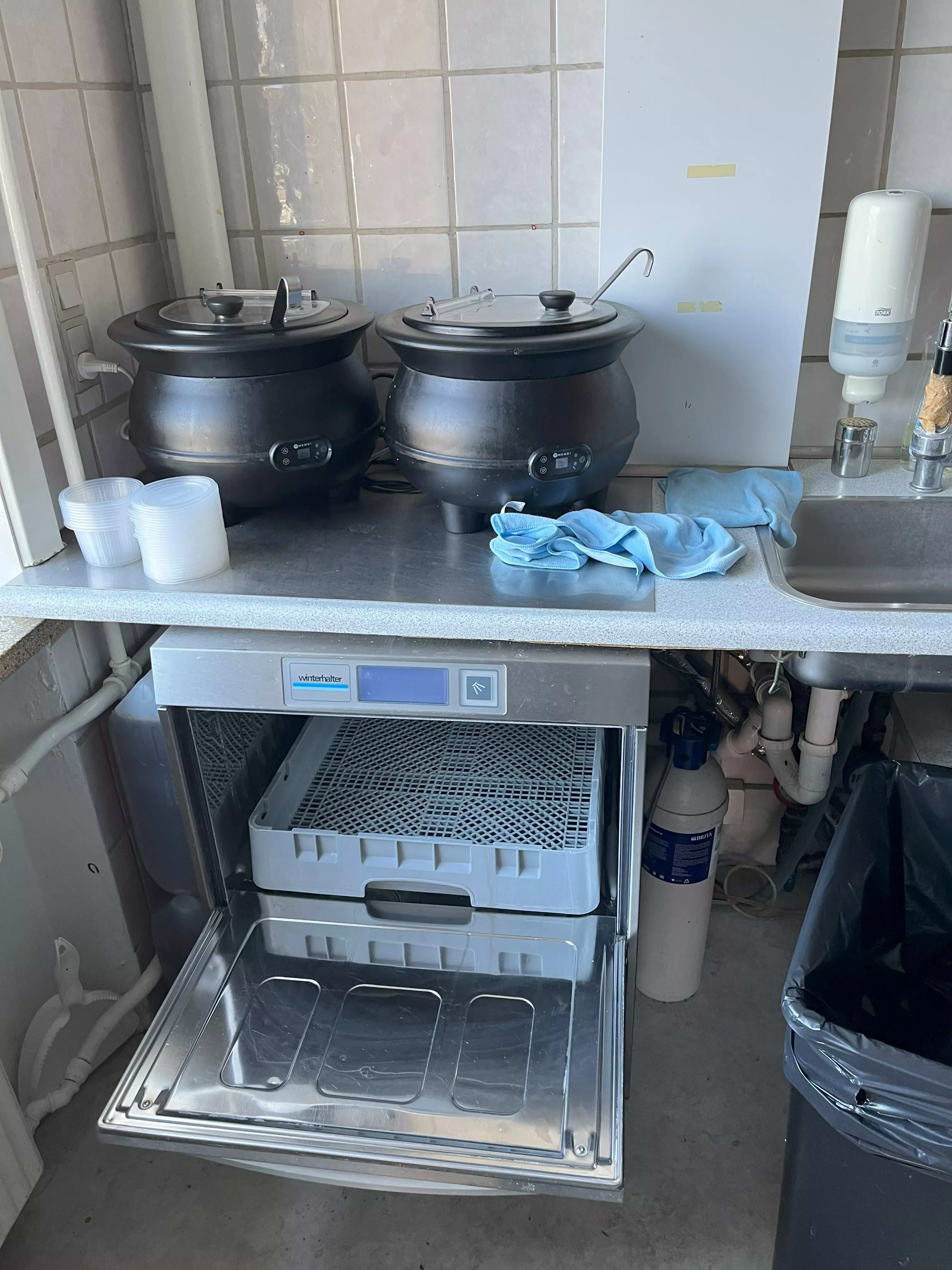 Køkken med to store gryder med låg og flere plastbeholdere. Et stålapparat med åben dør og et vask og skraldspande.