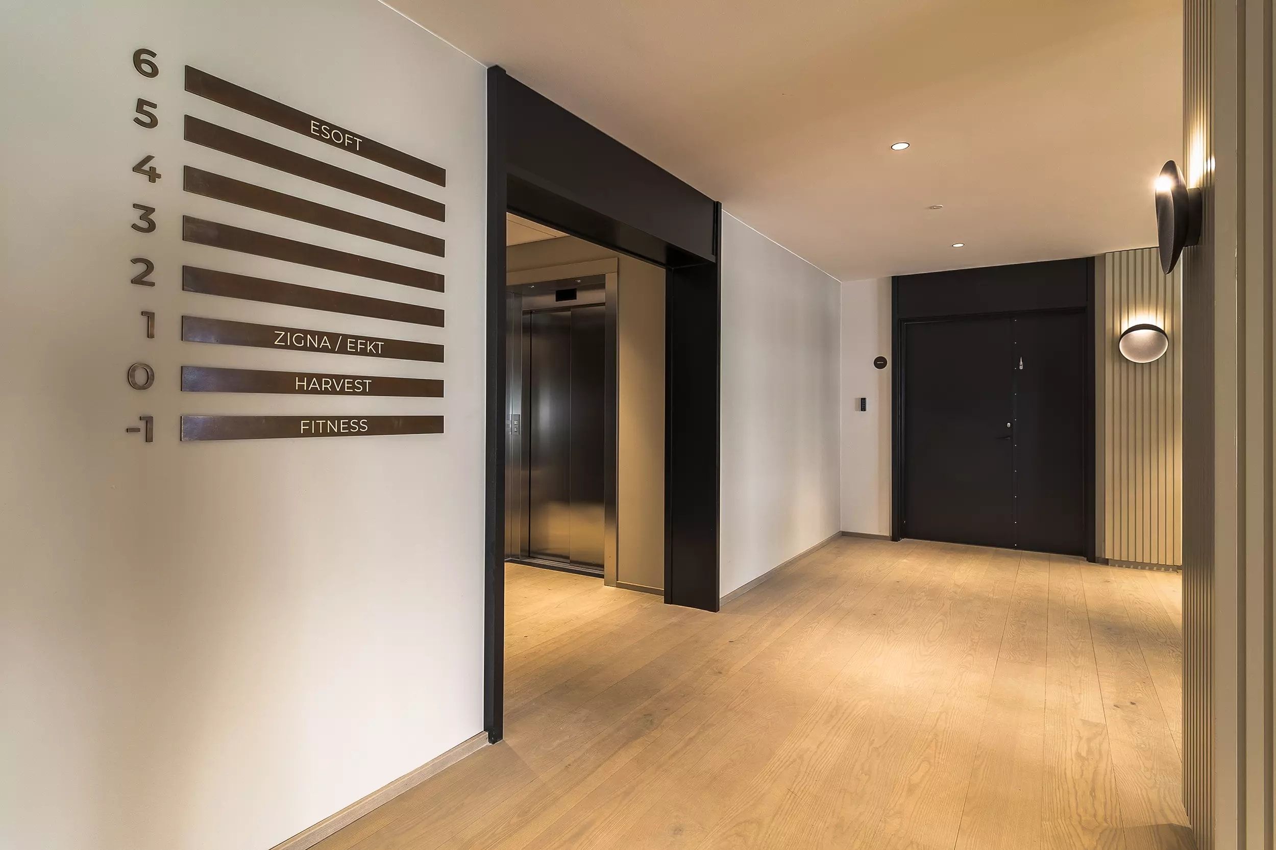 Indgangsparti til en bygning med loftsventilatorer, to elevatorer, hvid væg med bronze farvede nummerplade samt to indgange
