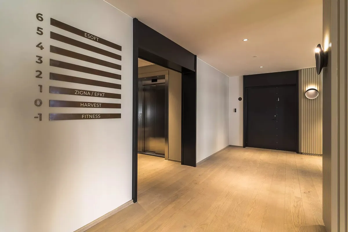 Indgangsparti til en bygning med loftsventilatorer, to elevatorer, hvid væg med bronze farvede nummerplade samt to indgange