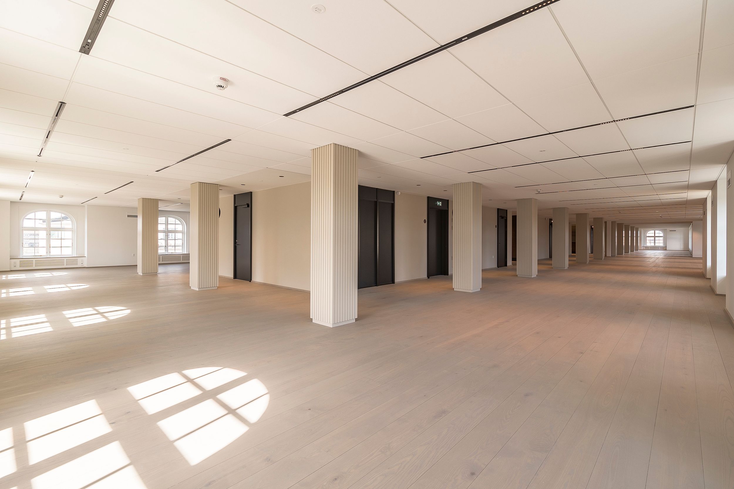 Elegant, rummelig kontorhall med højt loft, hvide vægge og lyse gulvplanker. Kolonnestilte glasdøre og vinduer giver masser