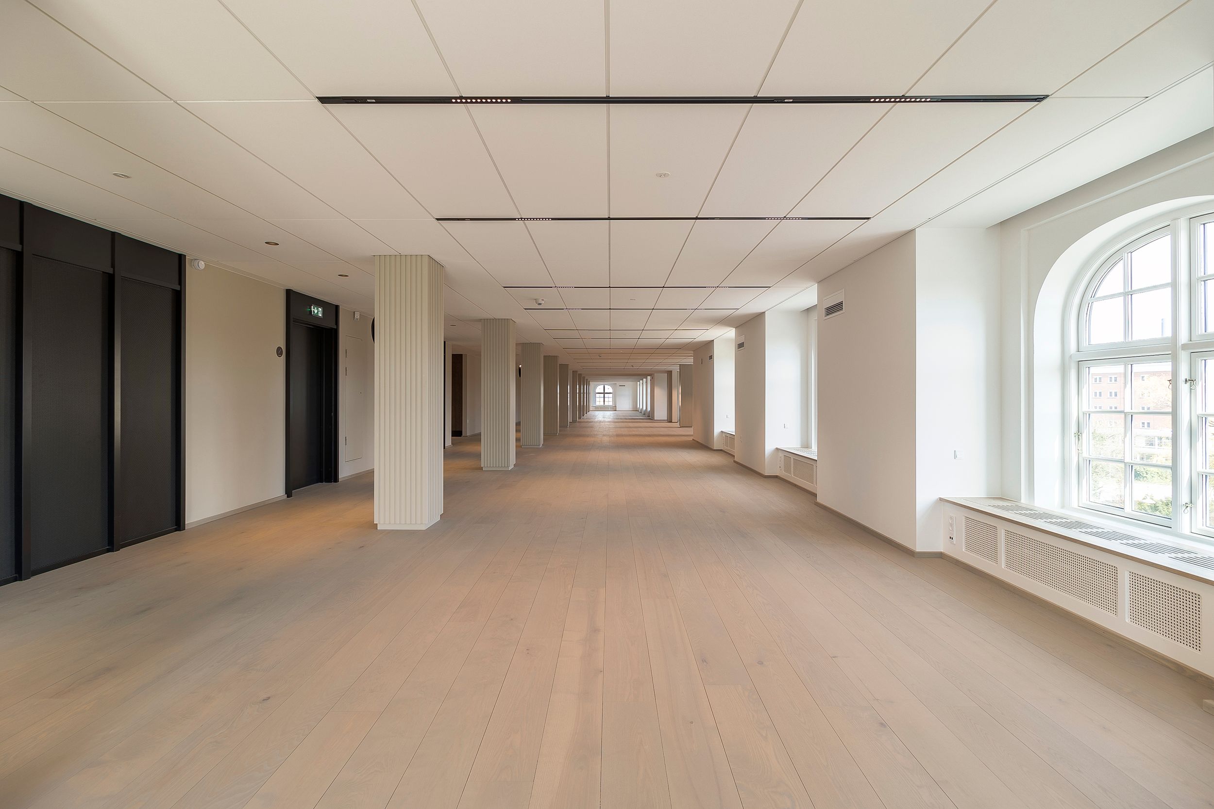 Langt og flot gange mellem to rækker søjler med hvid gulv og loft samt store runde vinduer med udsigt til bygning