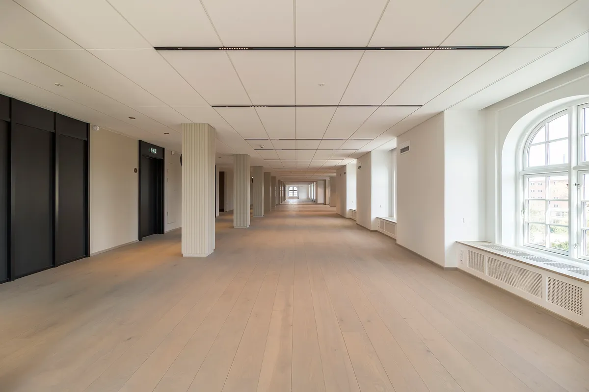 Langt og flot gange mellem to rækker søjler med hvid gulv og loft samt store runde vinduer med udsigt til bygning