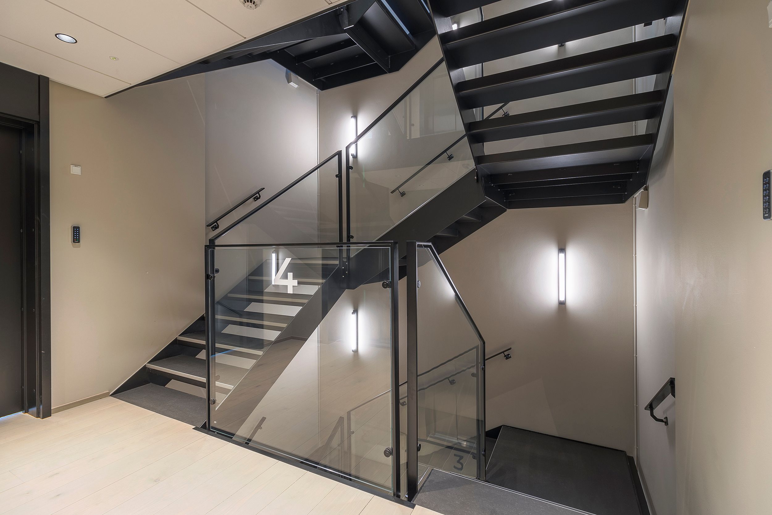 En moderne trappe med glasvægge og sort metal stiger op til et kontorlokal med nummeret 4. Styret i sort metal og gulvet med