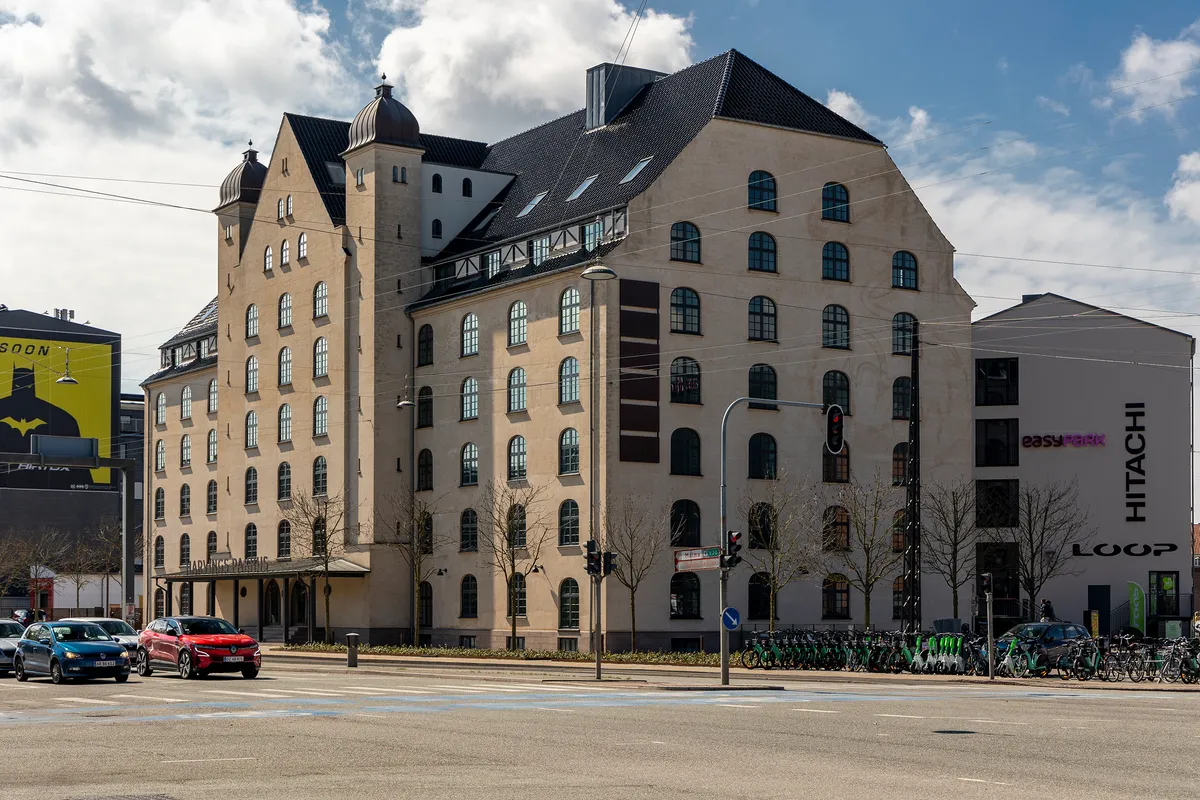 Bygning med flere vinduer, røde biler og cykler parkeret, en stor skilt med Hitachi Loop og biler kører på gaden