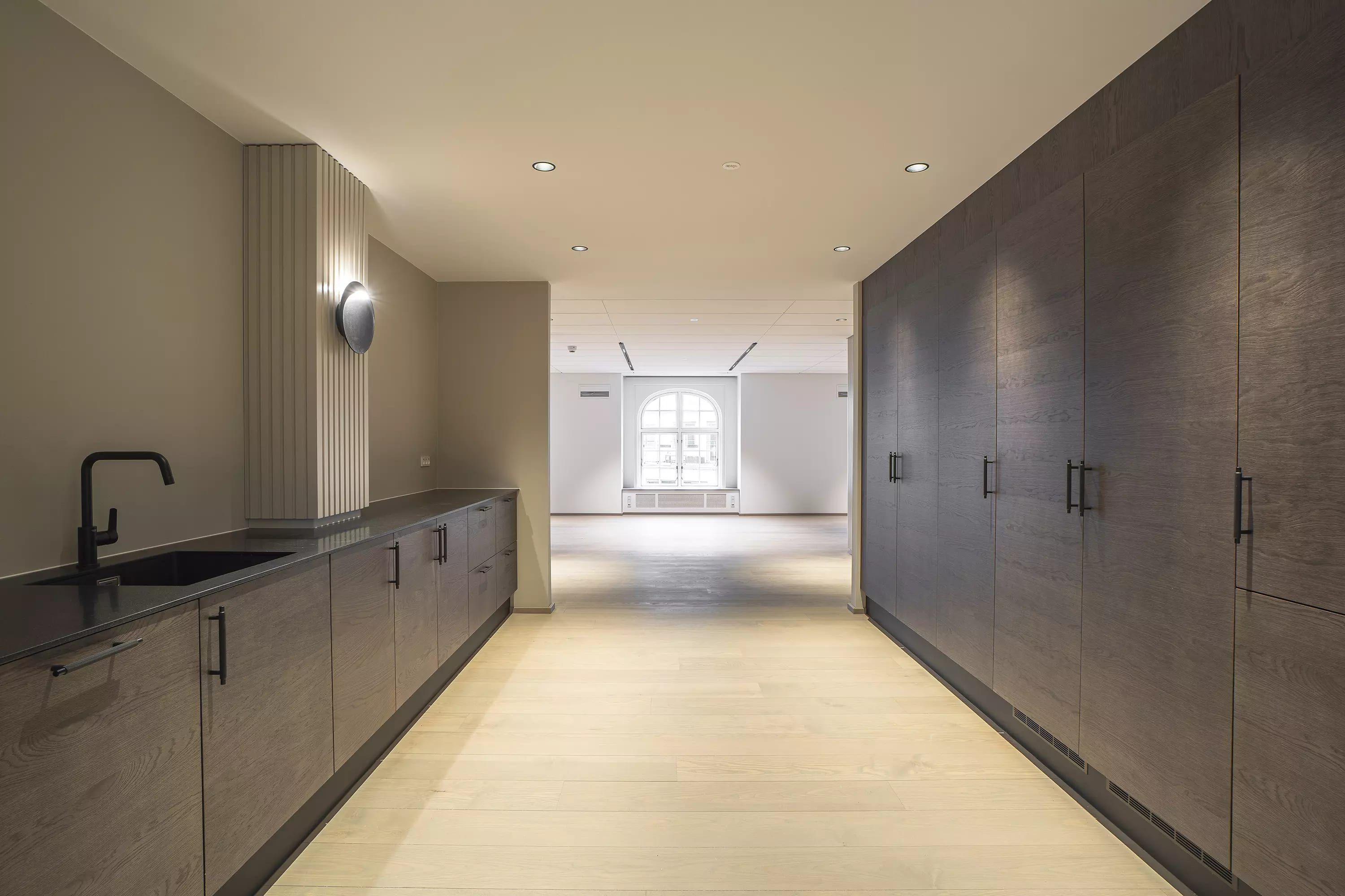 Minimalistisk køkken med lyse trægulve, store træskabe og moderne lysekroner. Sort vandhane og spejl i køkkenvæggen.