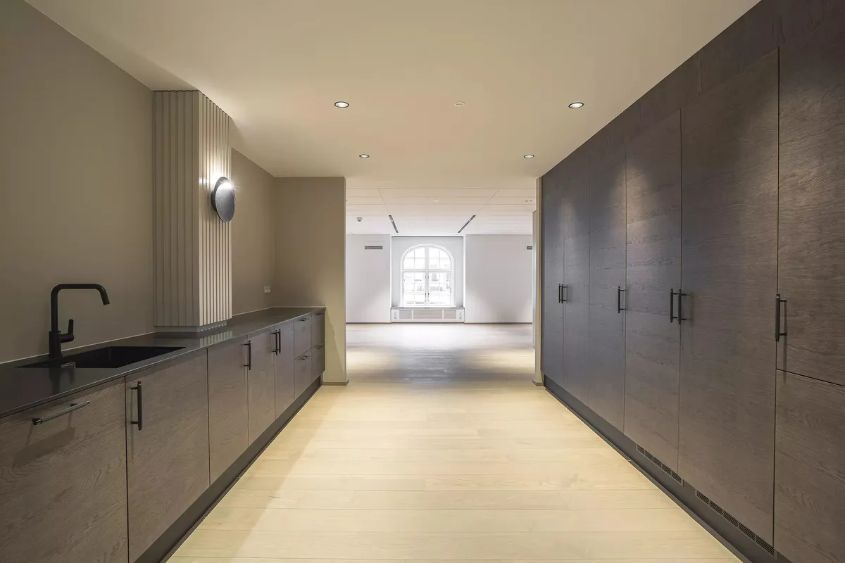 Minimalistisk køkken med lyse trægulve, store træskabe og moderne lysekroner. Sort vandhane og spejl i køkkenvæggen.