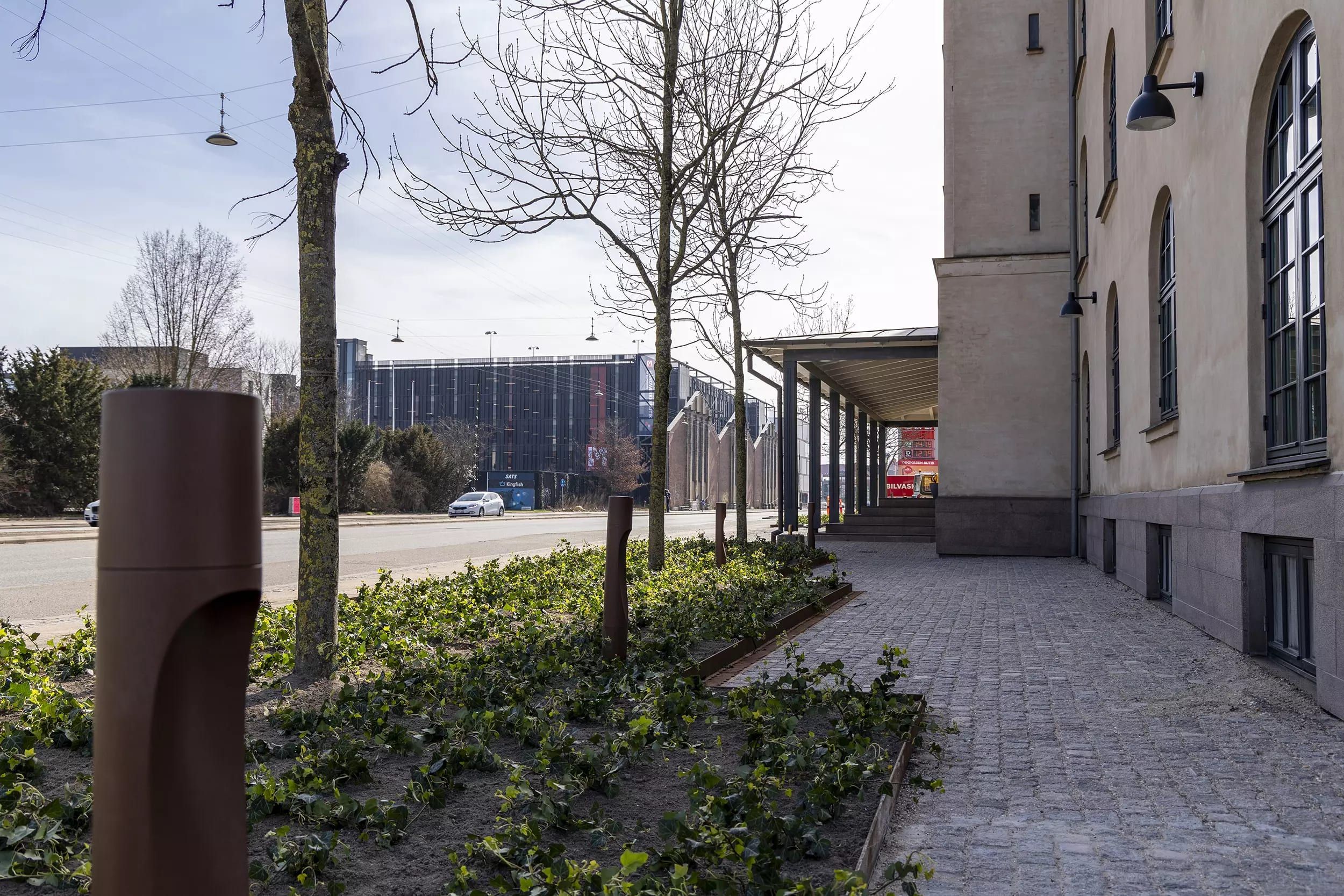 En gågade med terrasse, træer og buske, en bil og en bygning med teksten "Bilvasi"