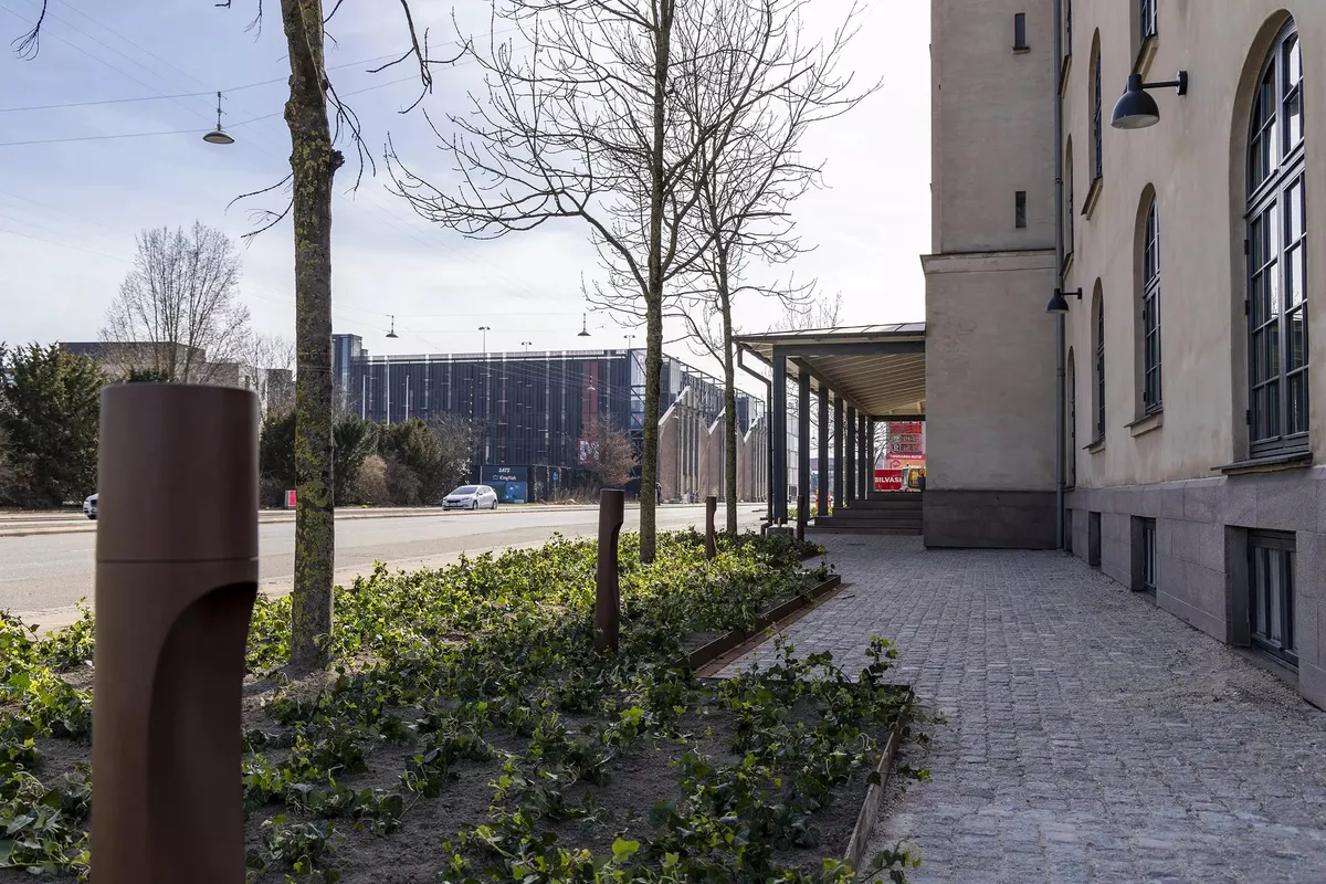 En gågade med terrasse, træer og buske, en bil og en bygning med teksten "Bilvasi"