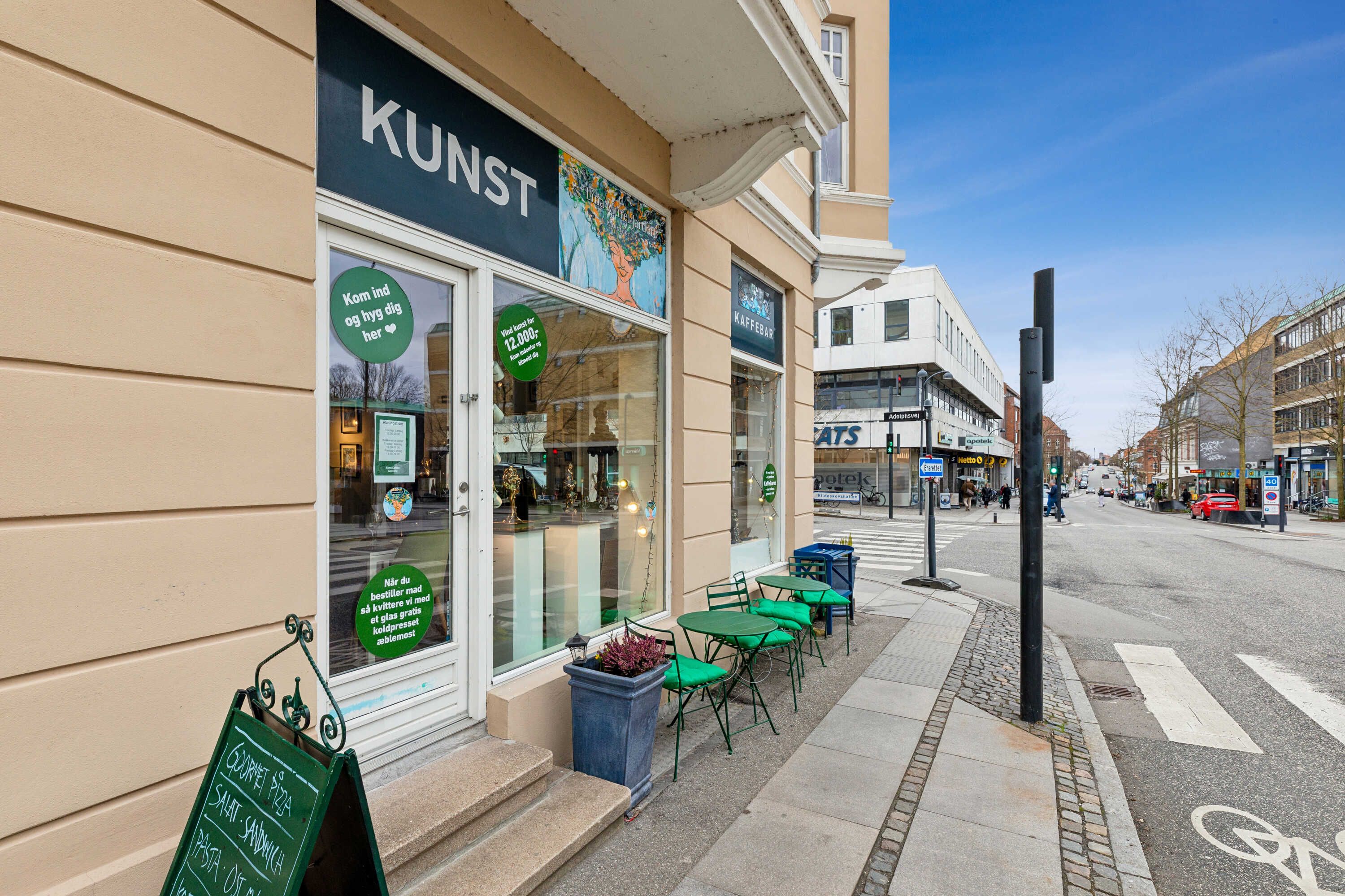 Kunst kafe ligger på Gentoftegade 33, og det er åbent for offentligheden. Der er grønne stole og borde udenfor caféen