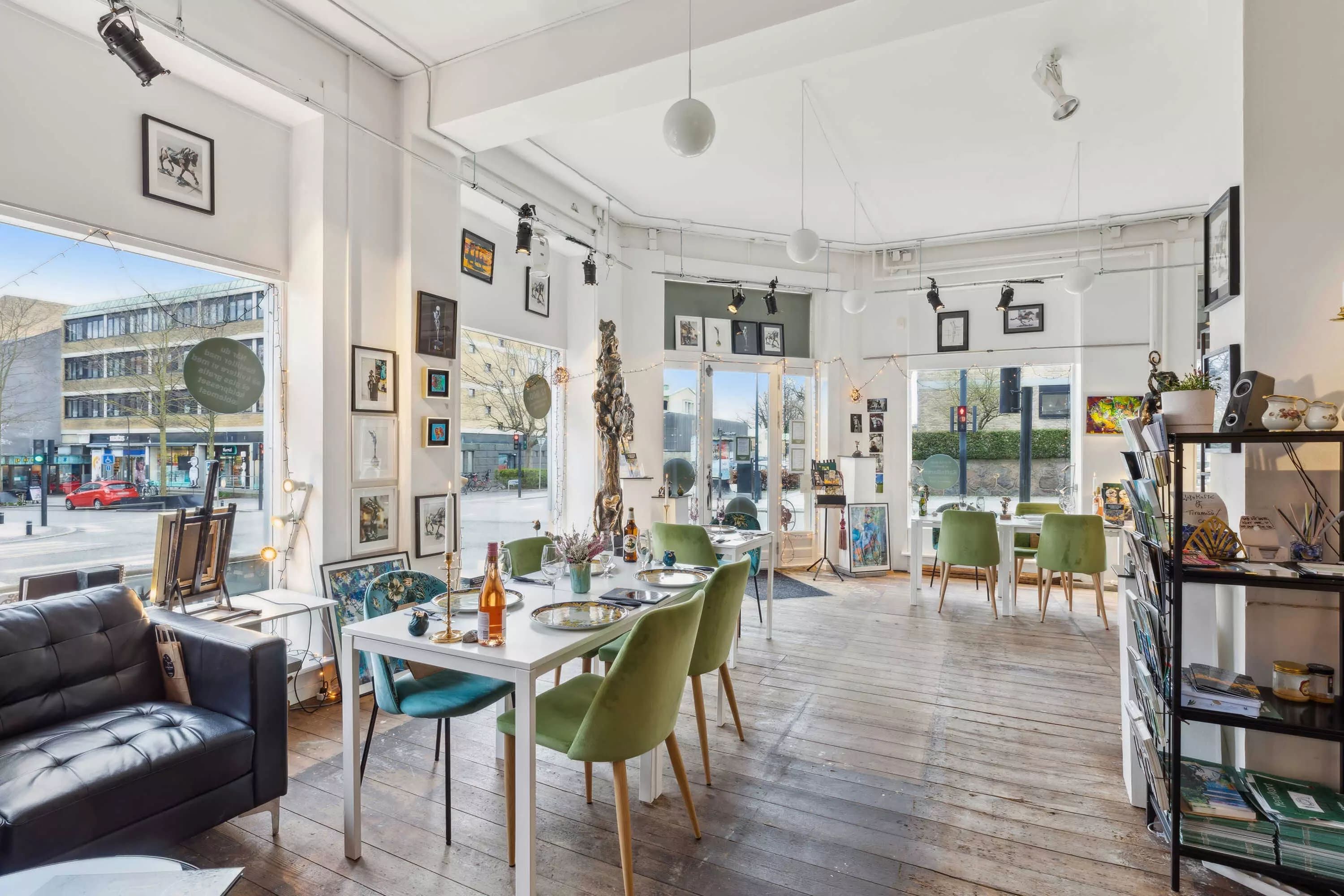 Inde i en elegant indretning med højloft, hvid væg og trægulv er der lyse grønne stole omkring borde, og et sofaset i sort.