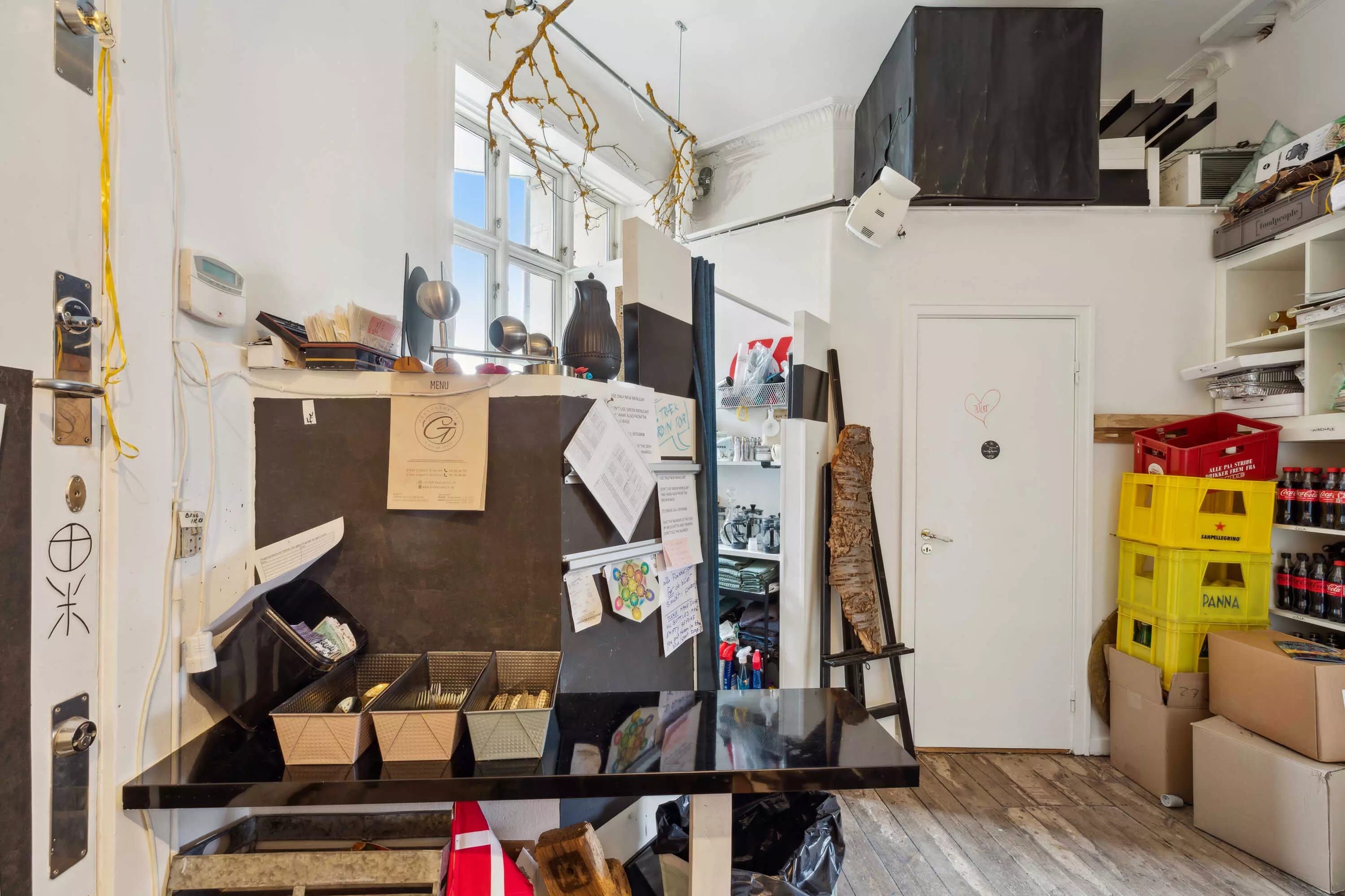 Et køkken med en bordplade, en køleskab og en loftsboks i loftet. Der er flere kasser og flasker, og et gulv med