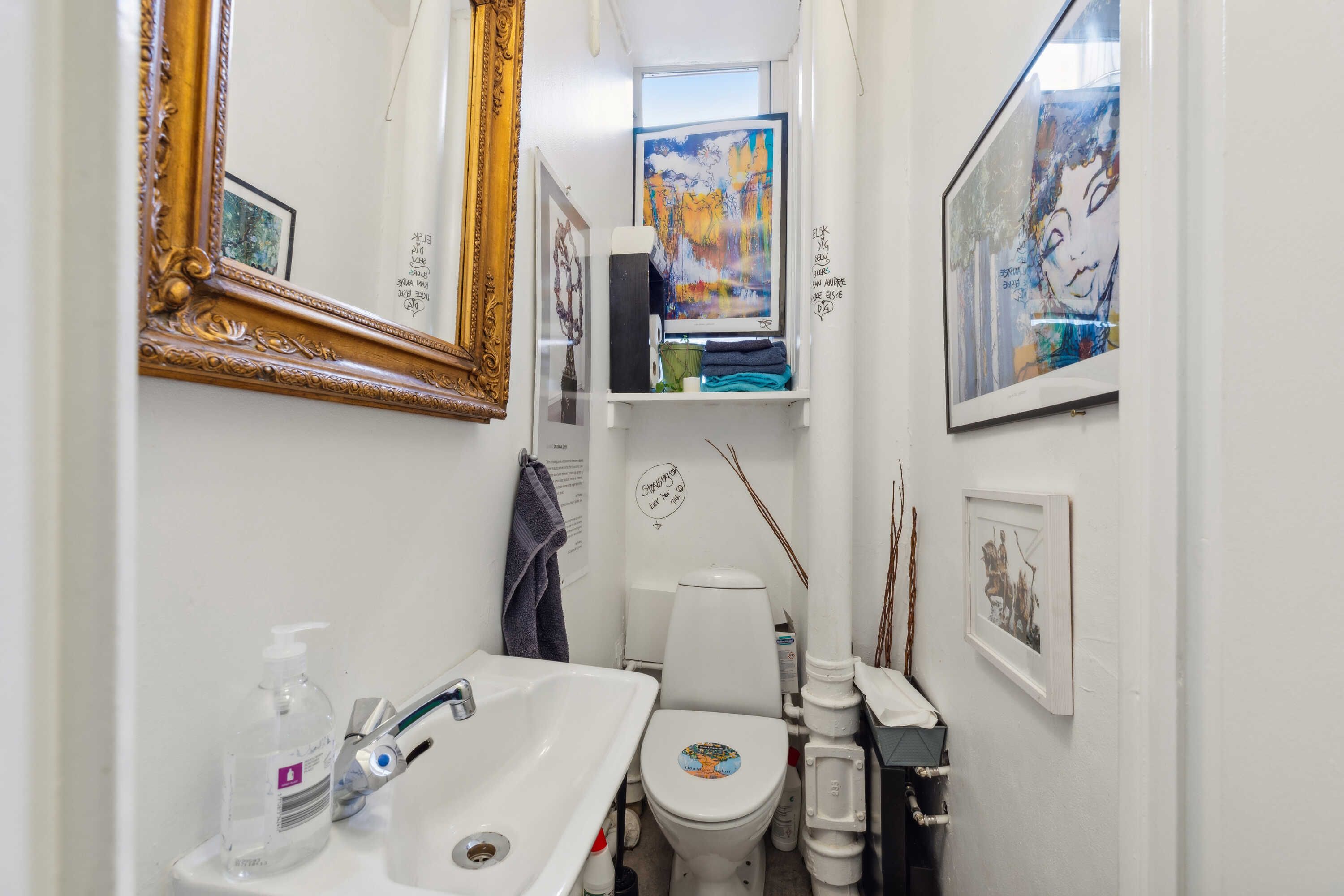 En lille toiletrum med hvidt toilet, håndvask og spejl samt flere billeder på væggen
