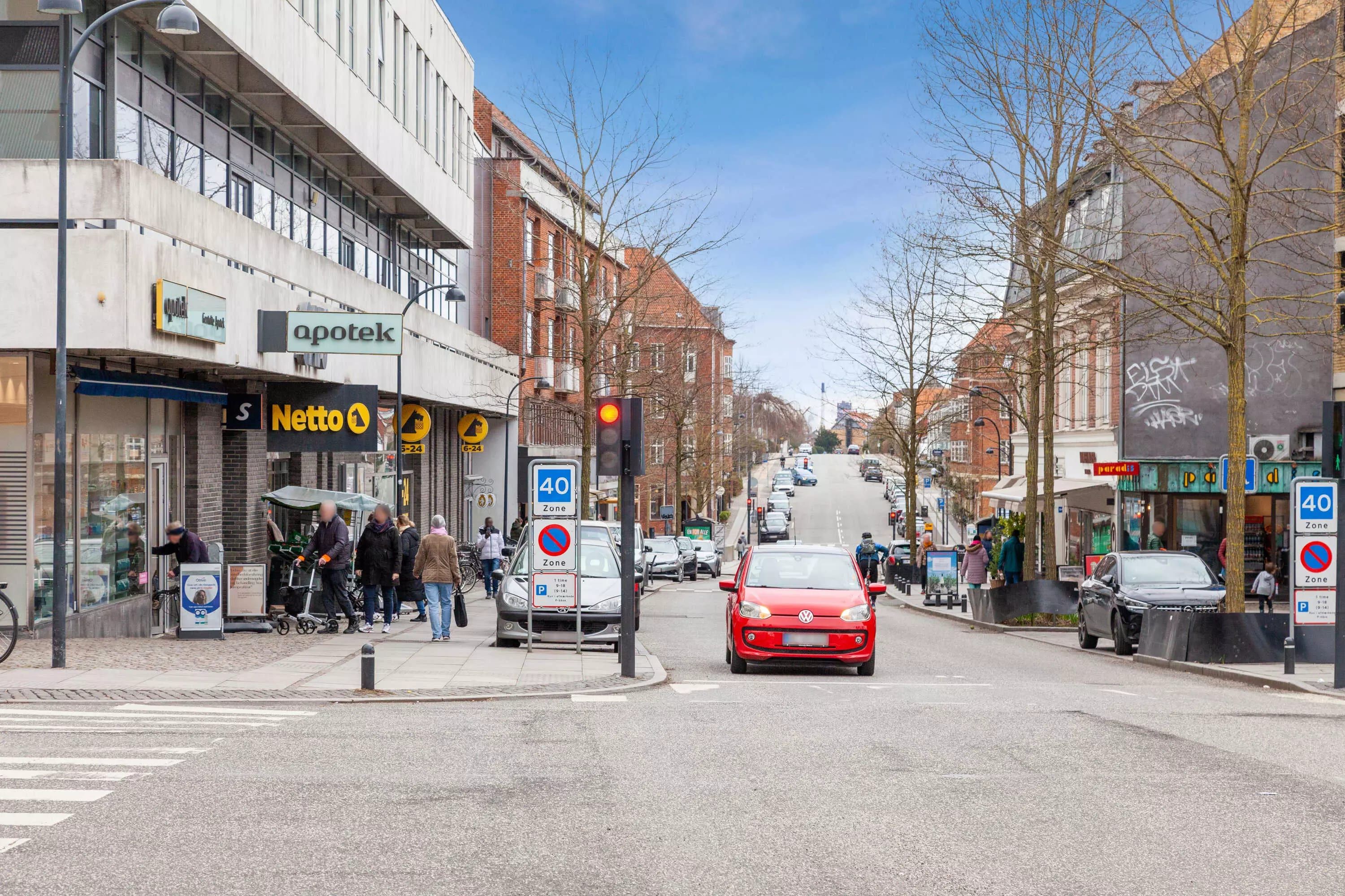 Biler kører på Gentoftegade og red lys. Mennesker og vogne står på fortovet, og der er butikker og skraldespande langs gaden.