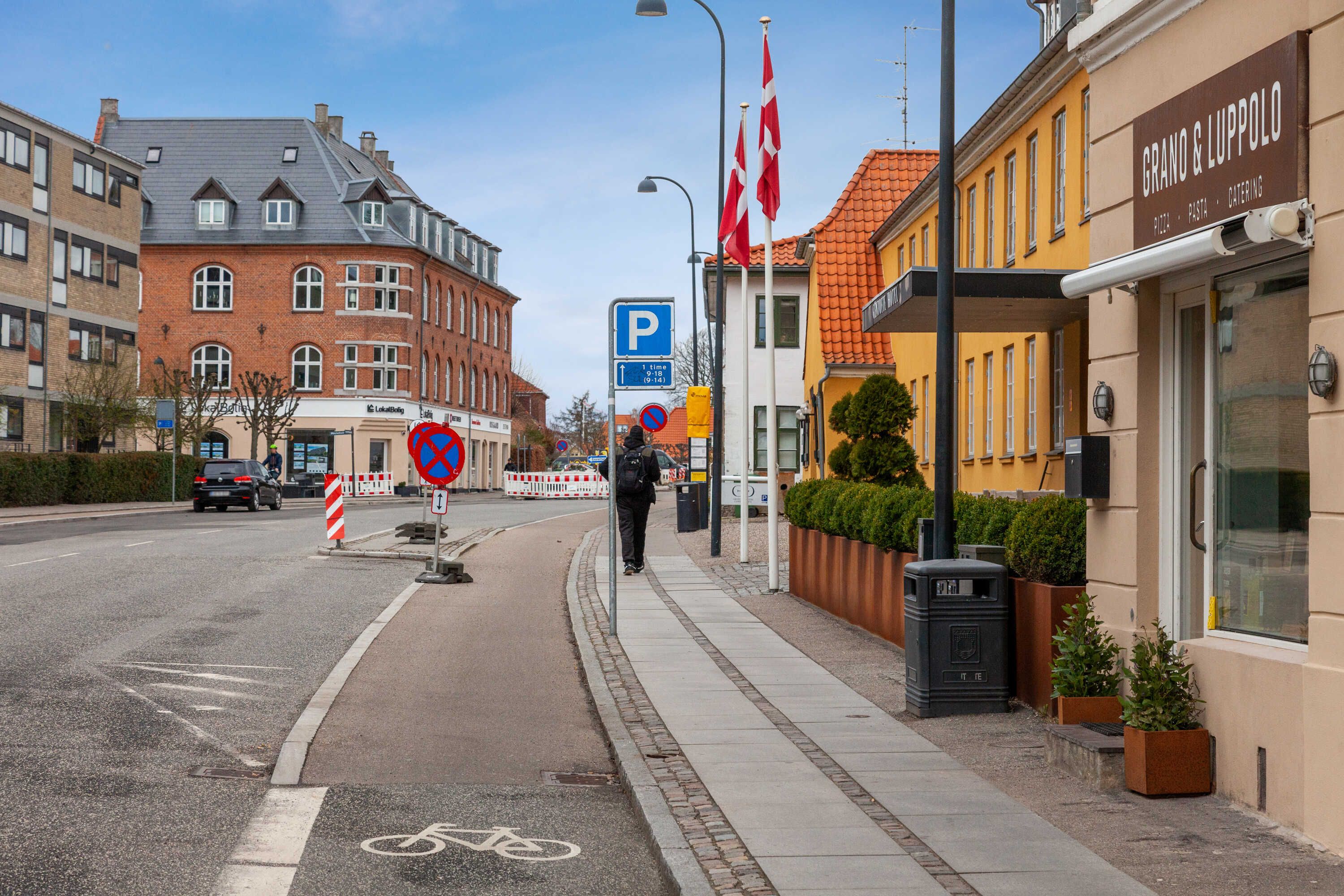 En mand går på fortov ved siden af et hotel med navnet Grano og Luppolo. Der er planter i potter og et skraldspand ved siden