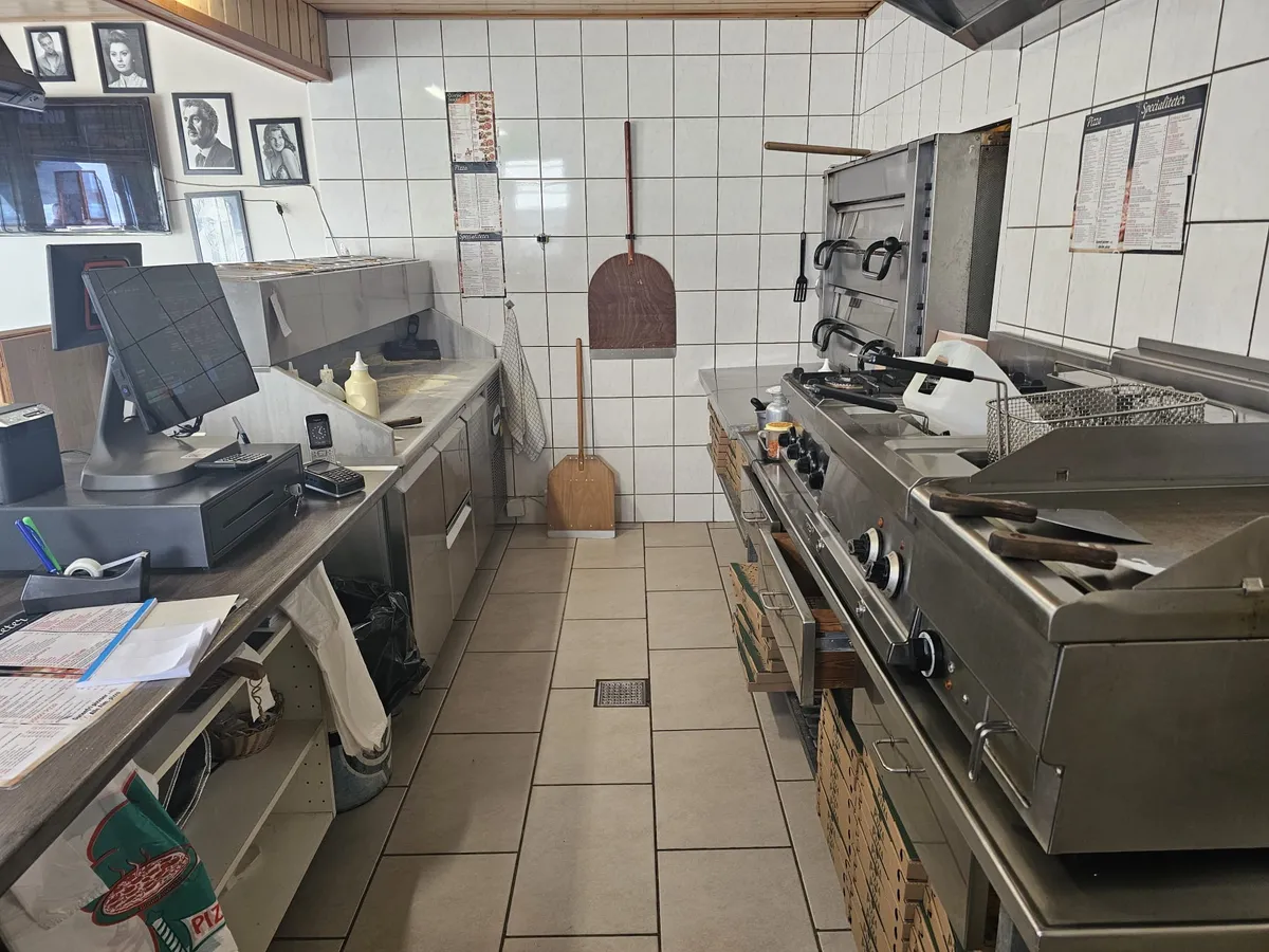 Køkken med en stor pizzaovn og en brændenælde, der er hængt op. Køkkenbord med computer og papirkurv samt flere tekstiler og