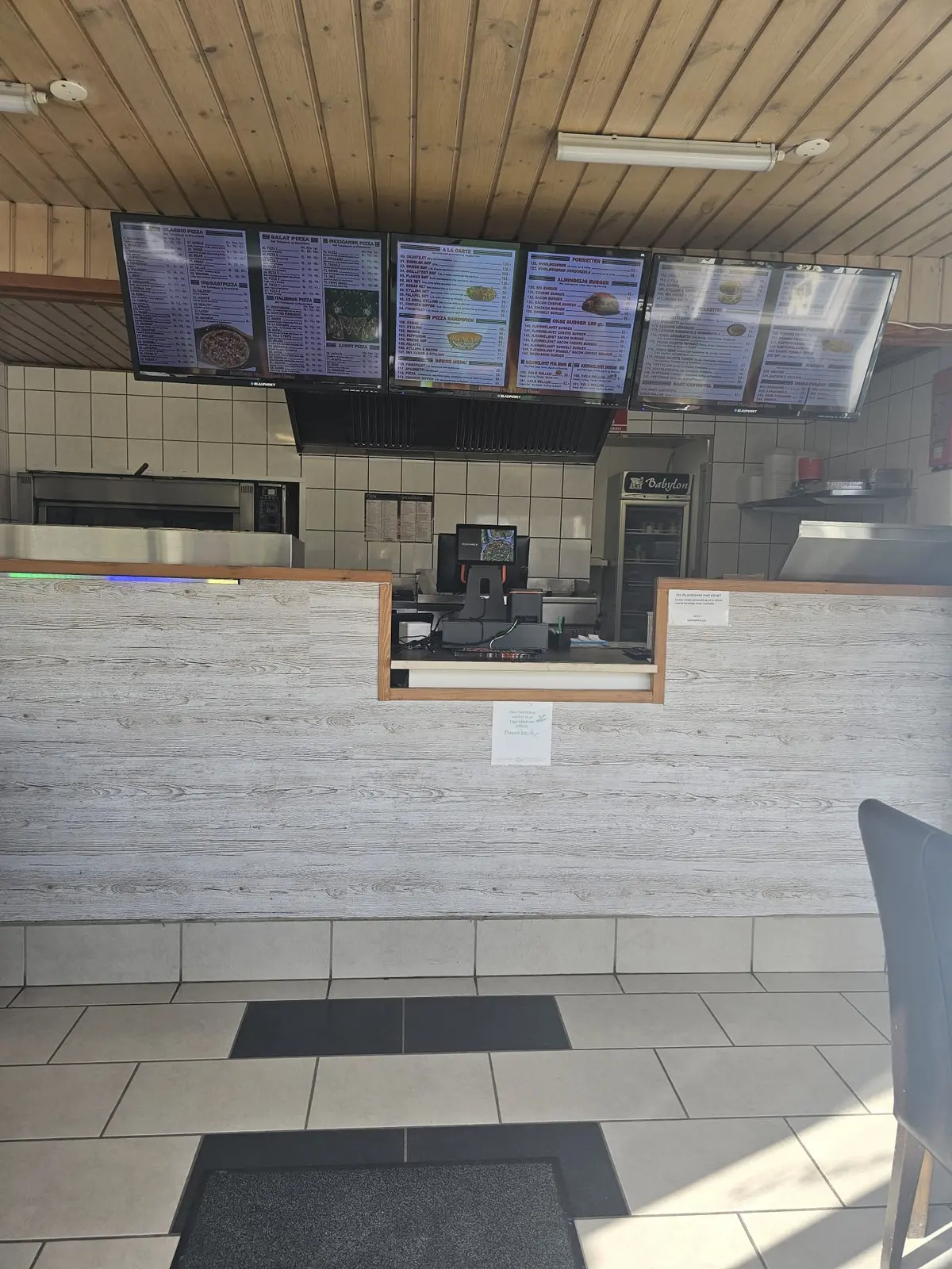 Menuer med pizza og burgere hænger på loftet i en pizzeria med en hvid og træagtig facade, en computer og et køleskab