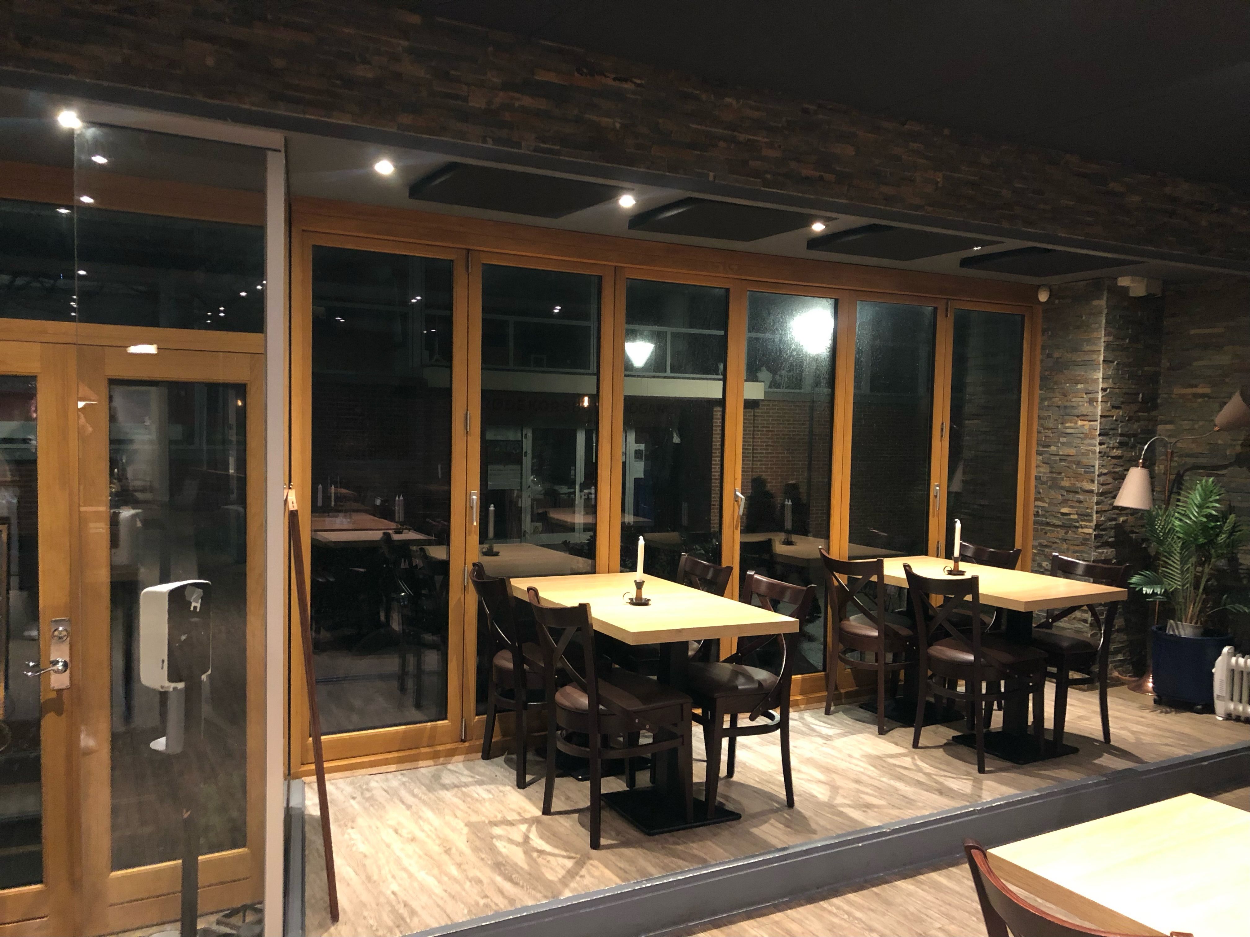 Indvendigt salgsrum med glasvægge, lysekroner, bord og stole, trægulv, en håndvask og en plante.