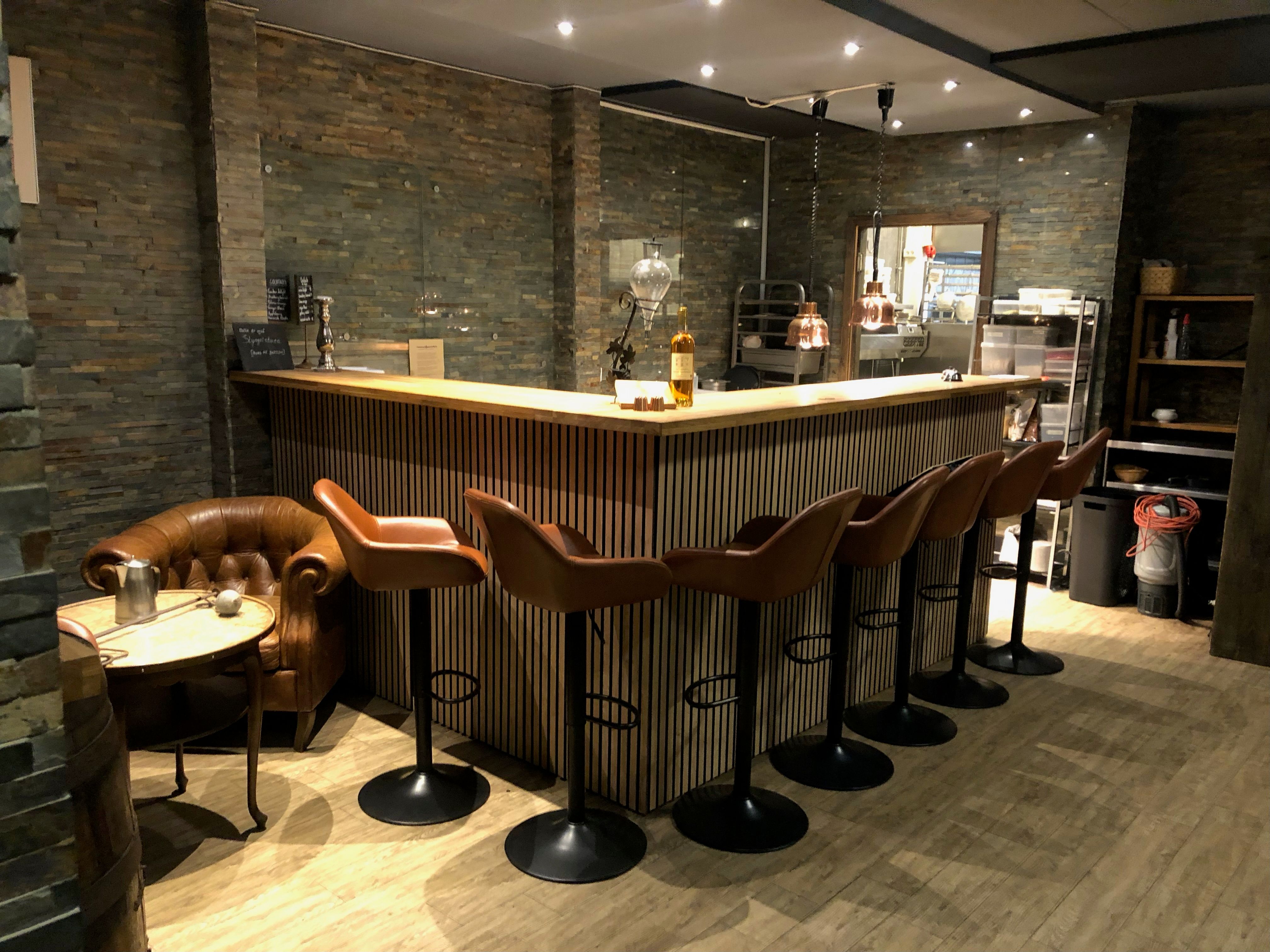 Et restaurantbord med barstole og en brændenælde. Der er en trægulv og en væg med sten. Der er en høj bænk med et glas og en