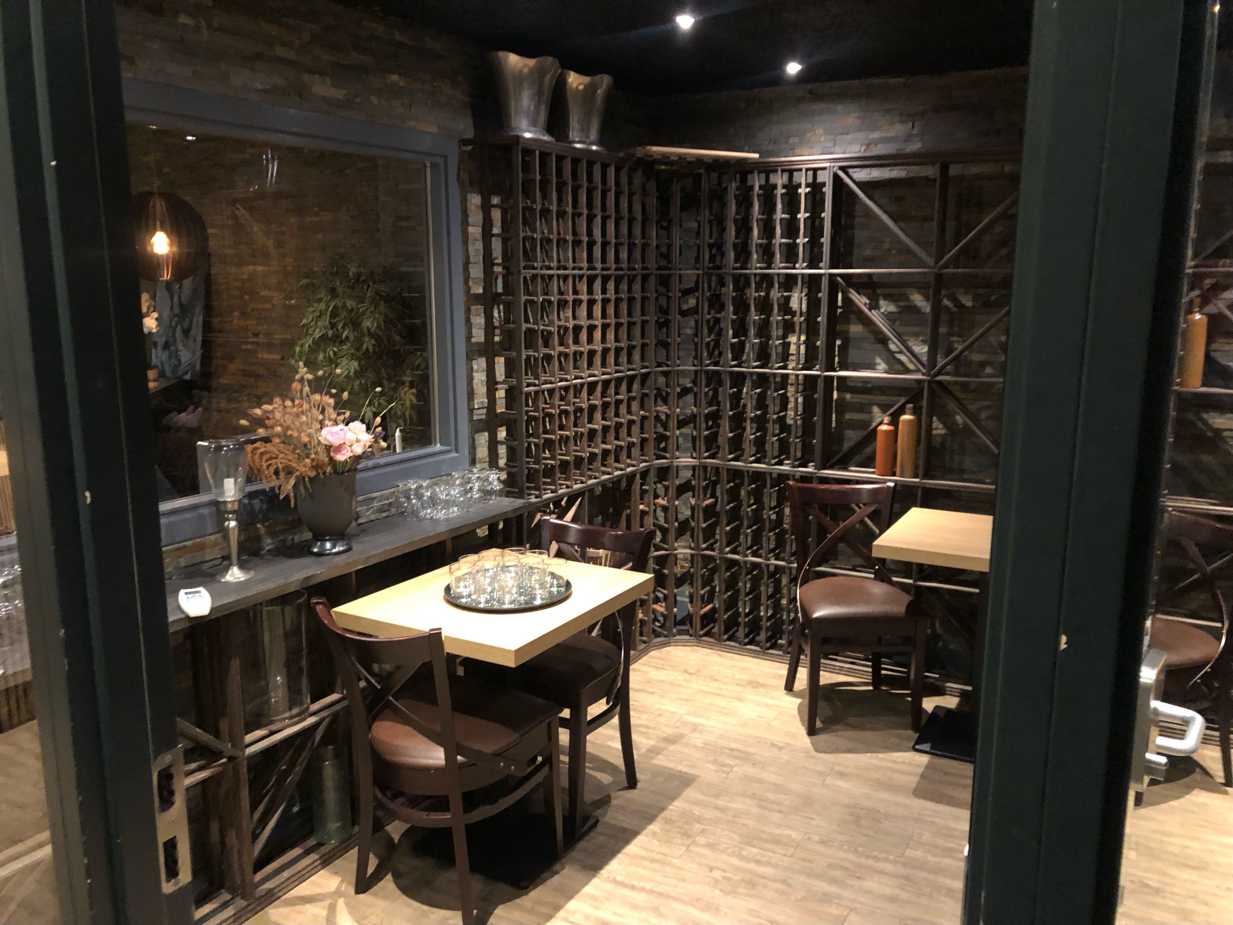 En lille vinbar med flere borde og stole samt vinreoler og en plante i en vase