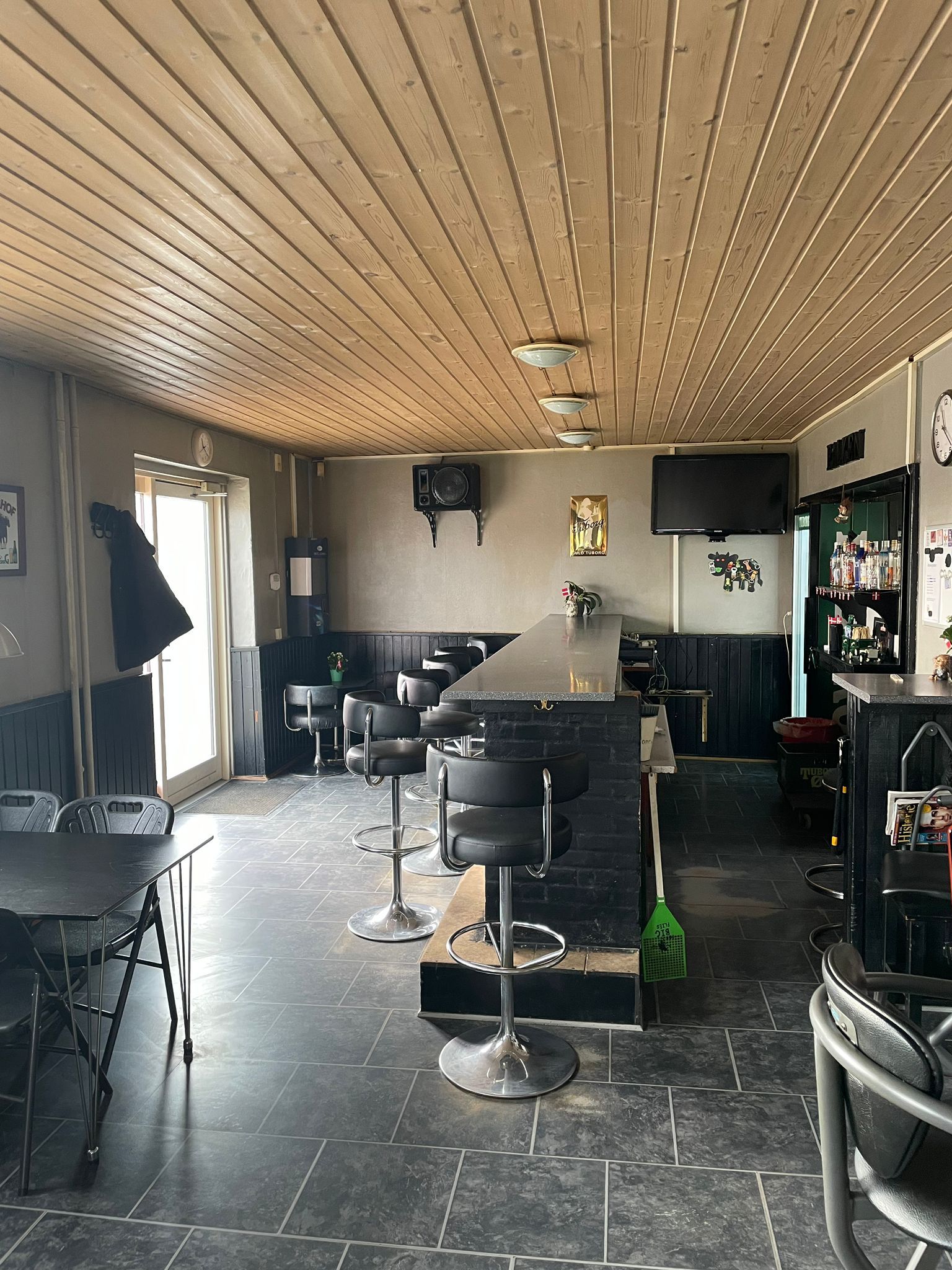 Indvendørs café med træloft, bar, stole, stolpe, træbord, TV, søjle, ølflaske, lysekroner, ure og skovl.