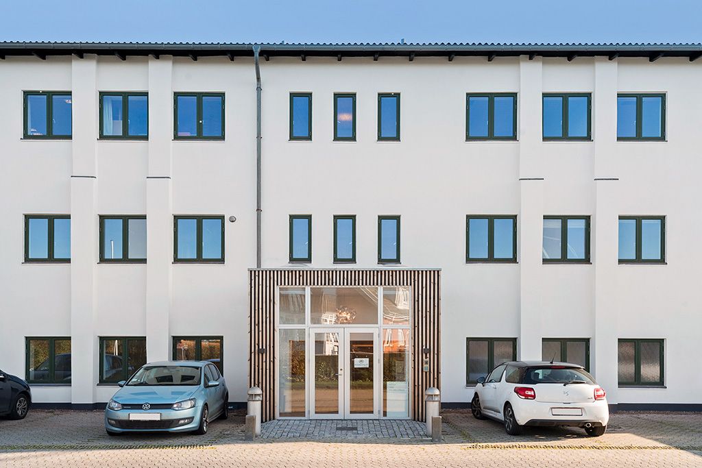 To parkerede biler, en Volkswagen og en Citroën, står foran en hvid bygning med mange vinduer og et glasdørsystem